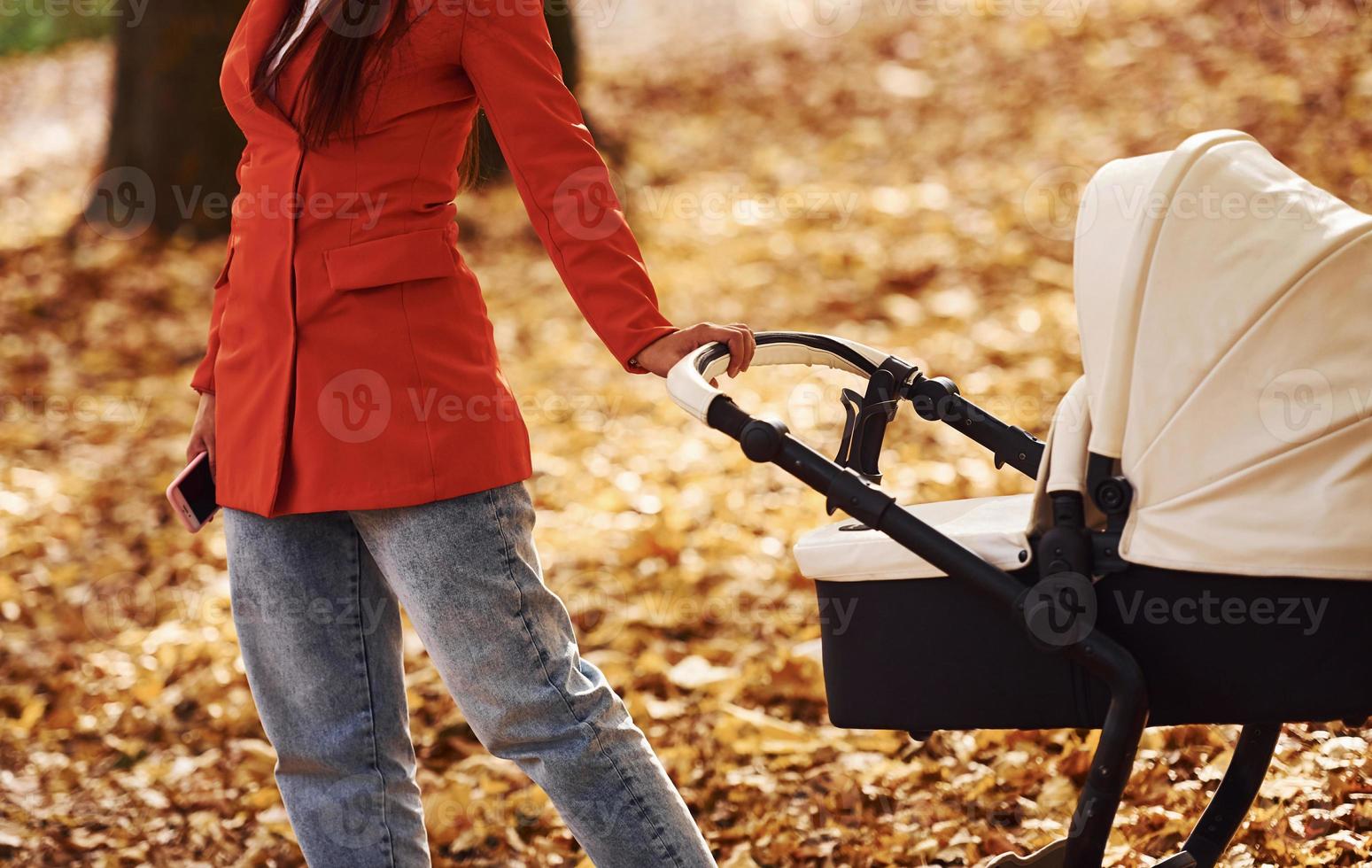 Close up view. Mother in red coat have a walk with her kid in the pram in the park at autumn time photo