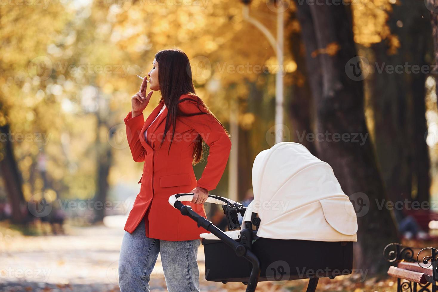 Mother in red coat have a walk with her kid in the pram in the park at autumn time and smoking photo
