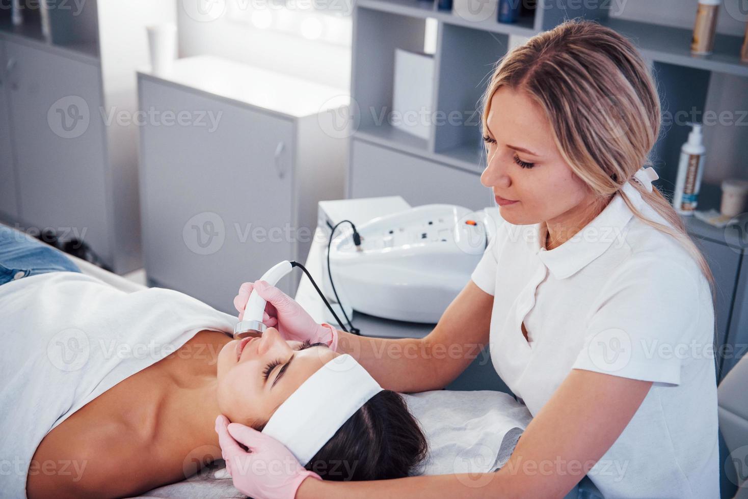 Woman lying down in spa salon and have face cleaning procedure by professional cosmetologist photo