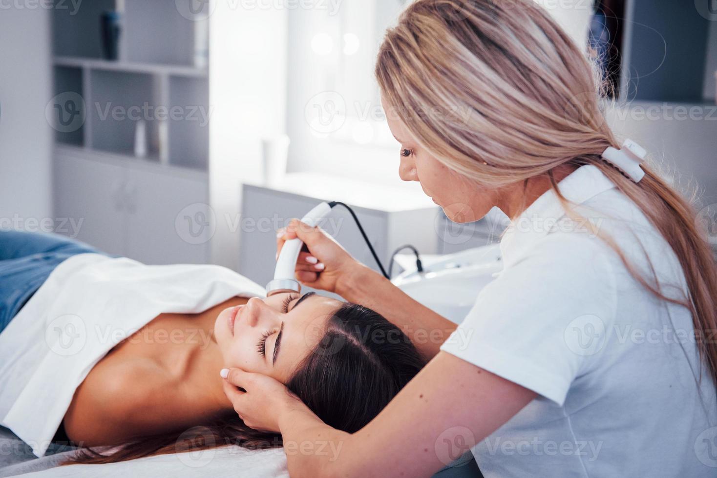 Woman lying down in spa salon and have face cleaning procedure by professional cosmetologist photo