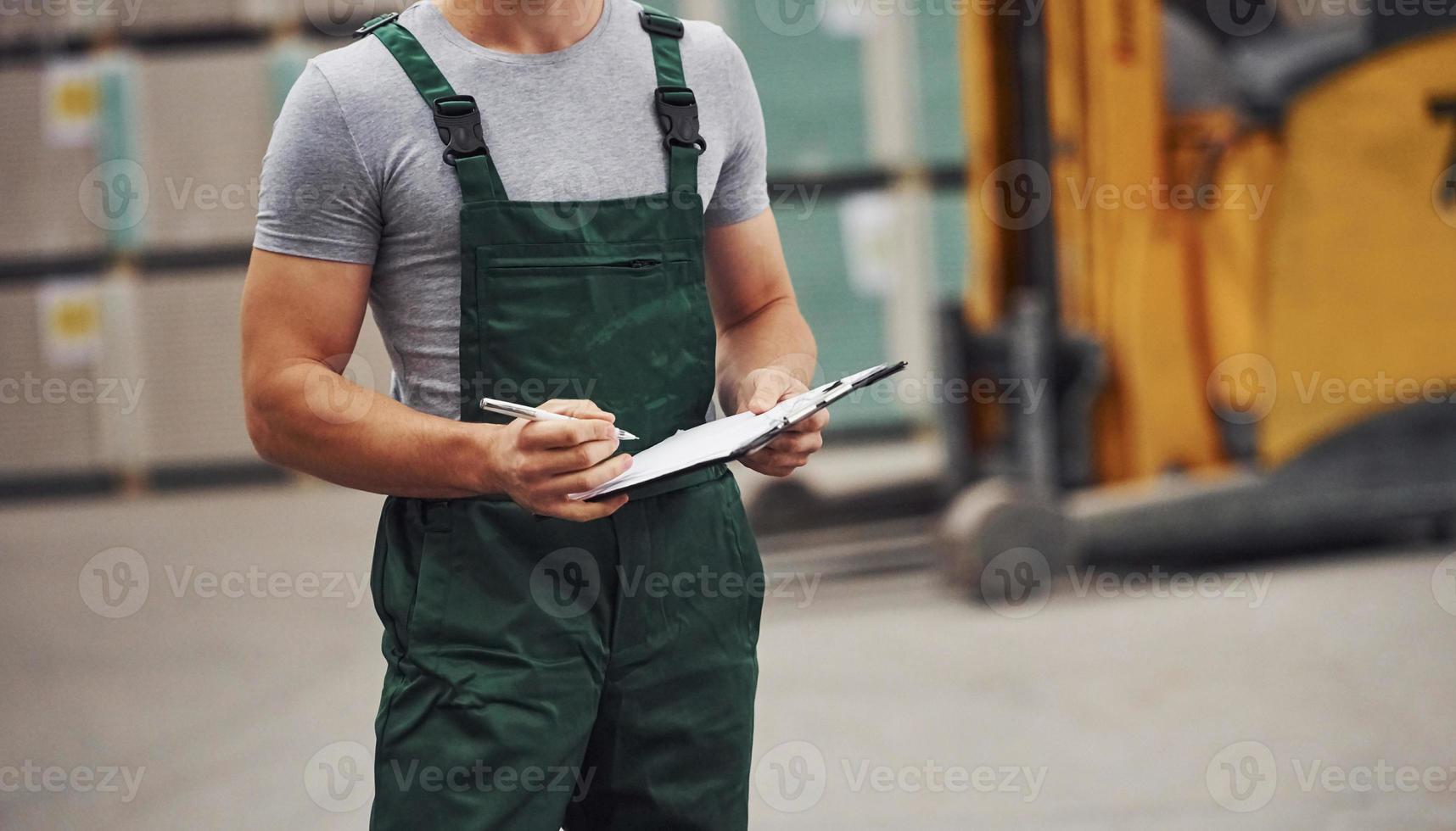 Storage worker in green colored uniform and notepad in hands checks production photo