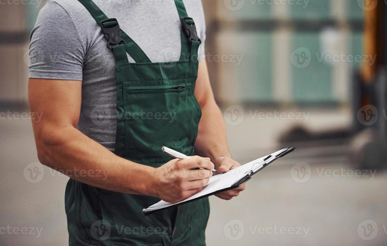 Storage worker in green colored uniform and notepad in hands checks production photo