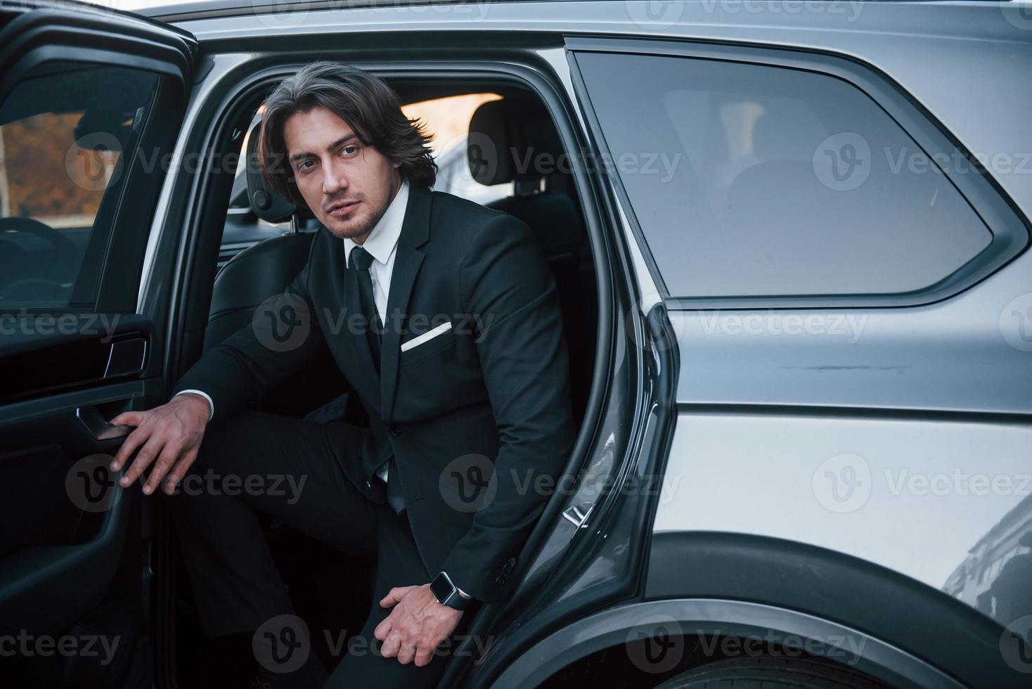Walks out from car. Young businessman in black suit and tie inside modern automobile photo