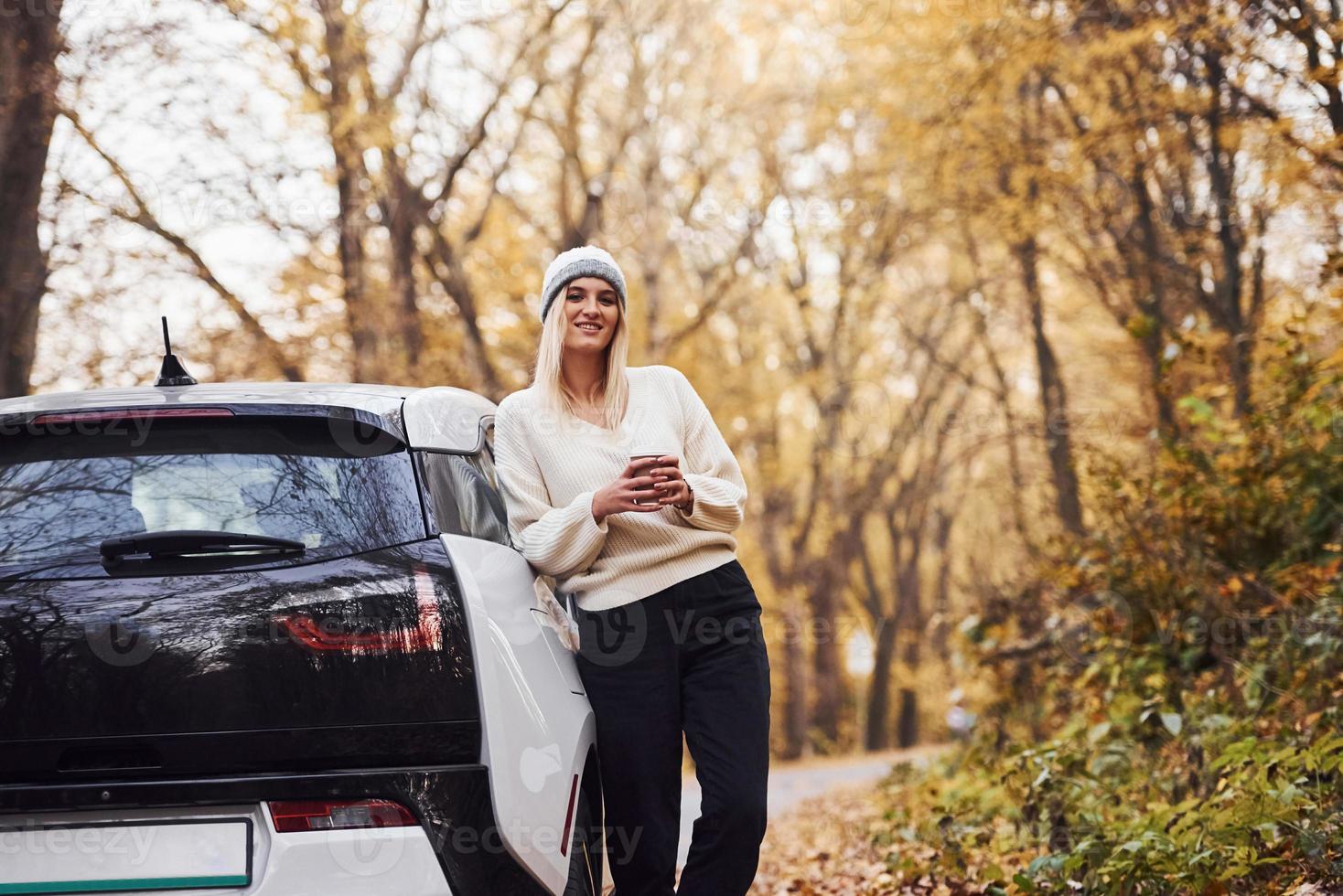 With cup of drink in hands. Girl have autumn trip by car. Modern brand new automobile in the forest photo