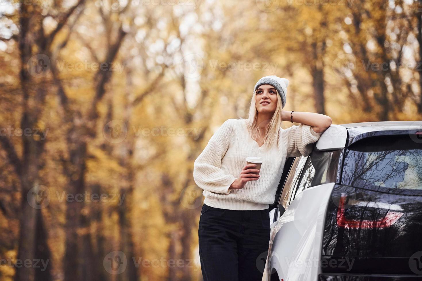 With cup of drink in hands. Girl have autumn trip by car. Modern brand new automobile in the forest photo