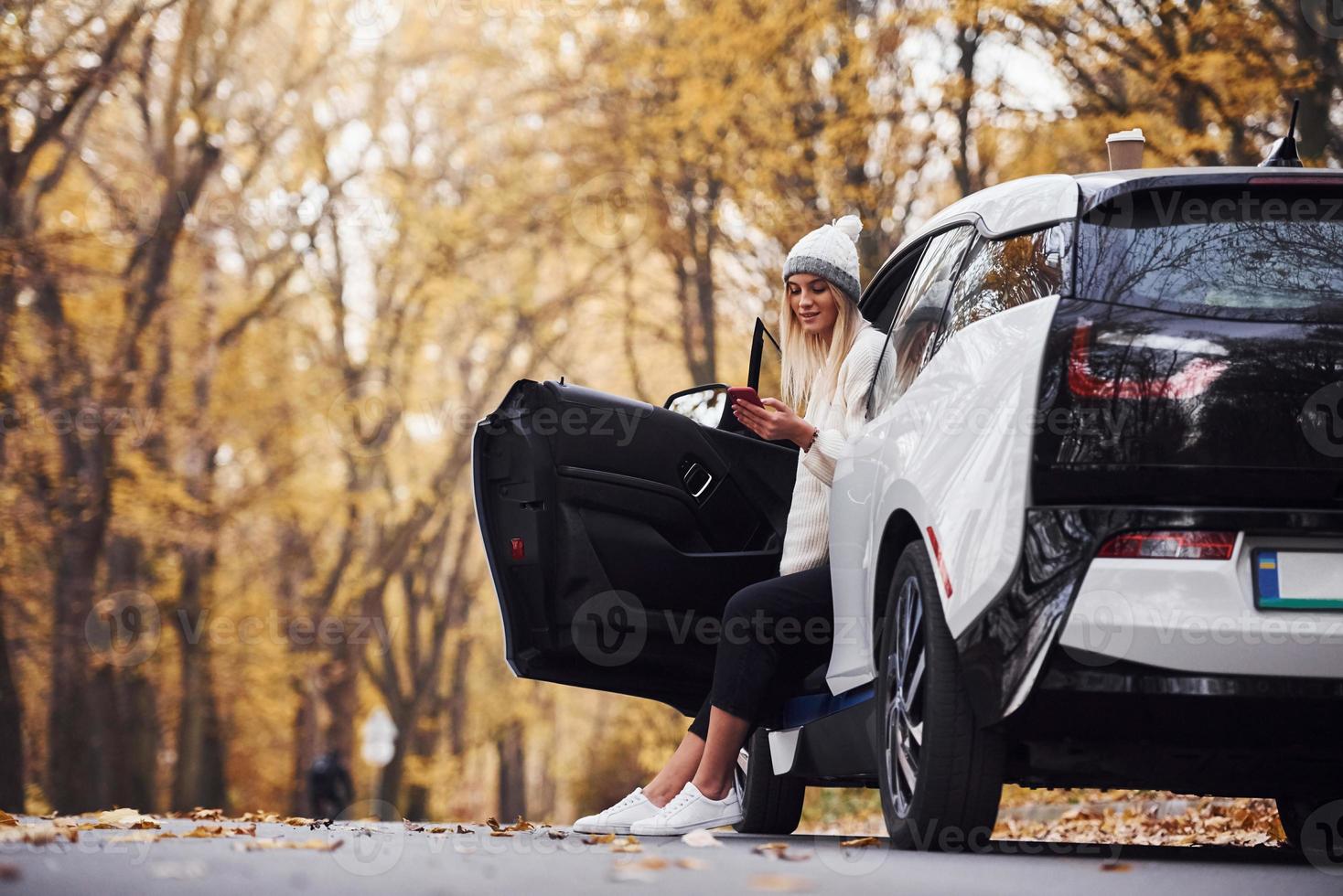 usando un teléfono inteligente. chica tiene viaje de otoño en coche. automóvil nuevo y moderno en el bosque foto