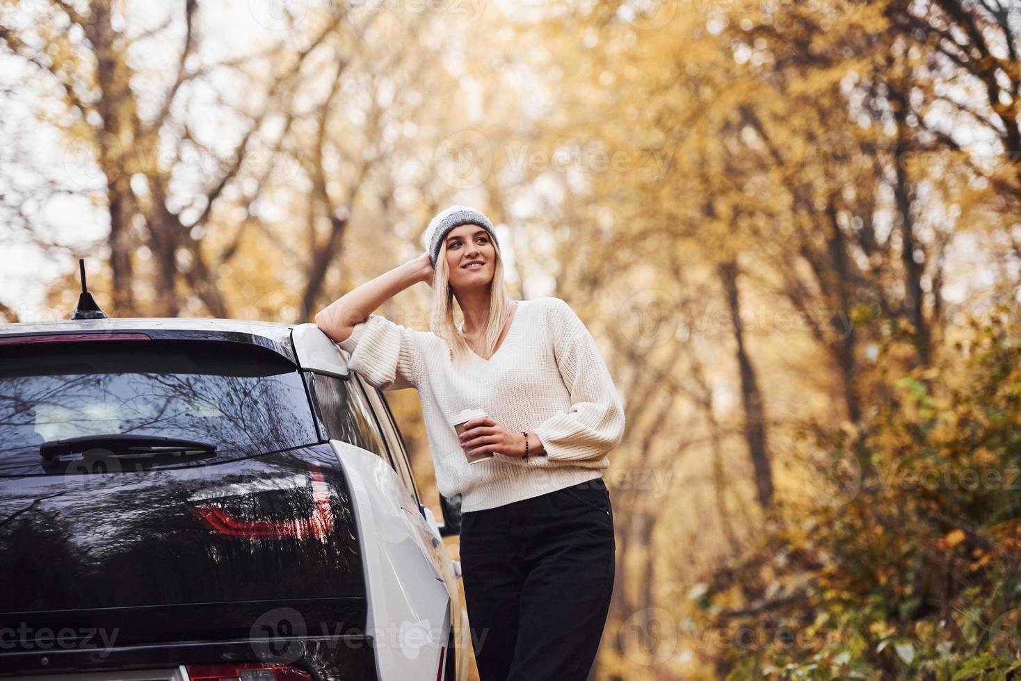 With cup of drink in hands. Girl have autumn trip by car. Modern brand new automobile in the forest photo