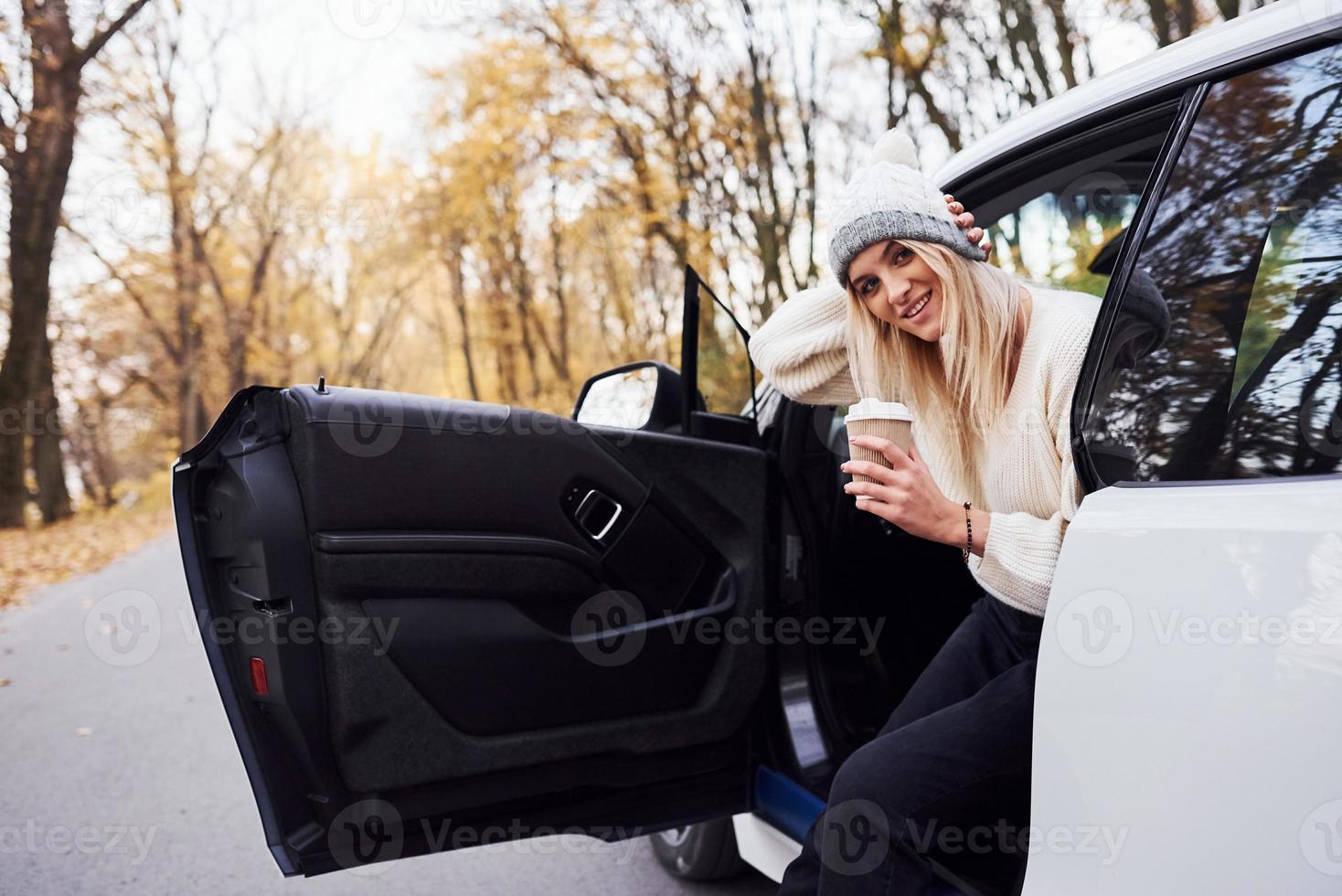 con taza de bebida en las manos. chica tiene viaje de otoño en coche. automóvil nuevo y moderno en el bosque foto