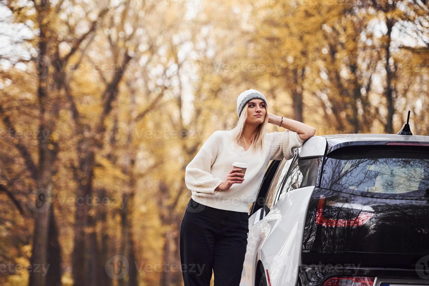 With cup of drink in hands. Girl have autumn trip by car. Modern brand new automobile in the forest photo