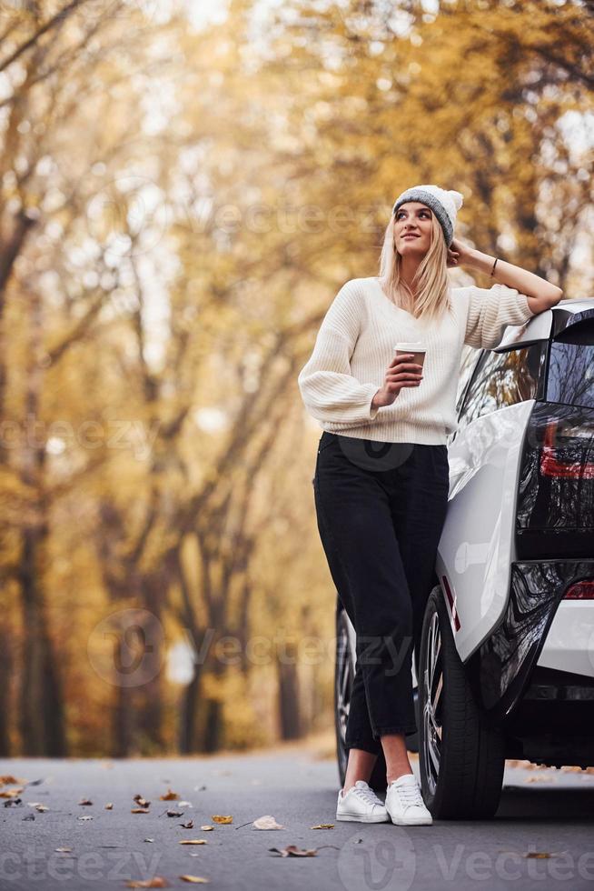 con taza de bebida en las manos. chica tiene viaje de otoño en coche. automóvil nuevo y moderno en el bosque foto