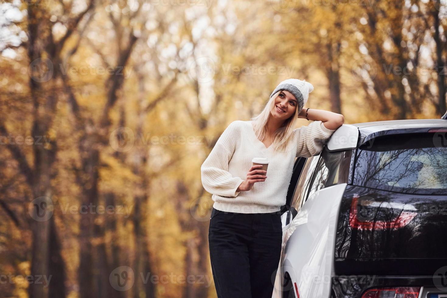 con taza de bebida en las manos. chica tiene viaje de otoño en coche. automóvil nuevo y moderno en el bosque foto