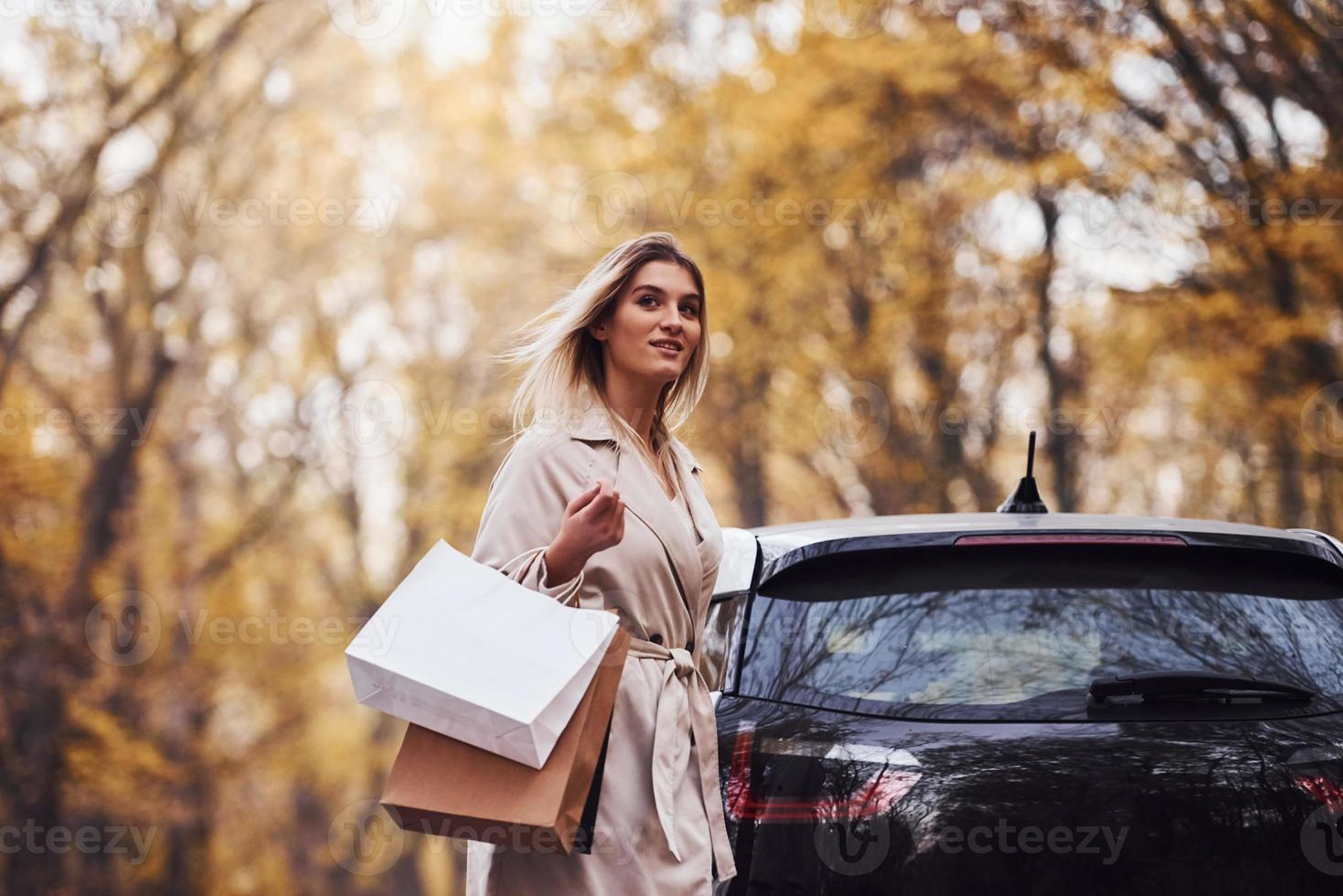 chica camina cerca del auto con bolsas de compras en las manos. automóvil nuevo y moderno en el bosque foto