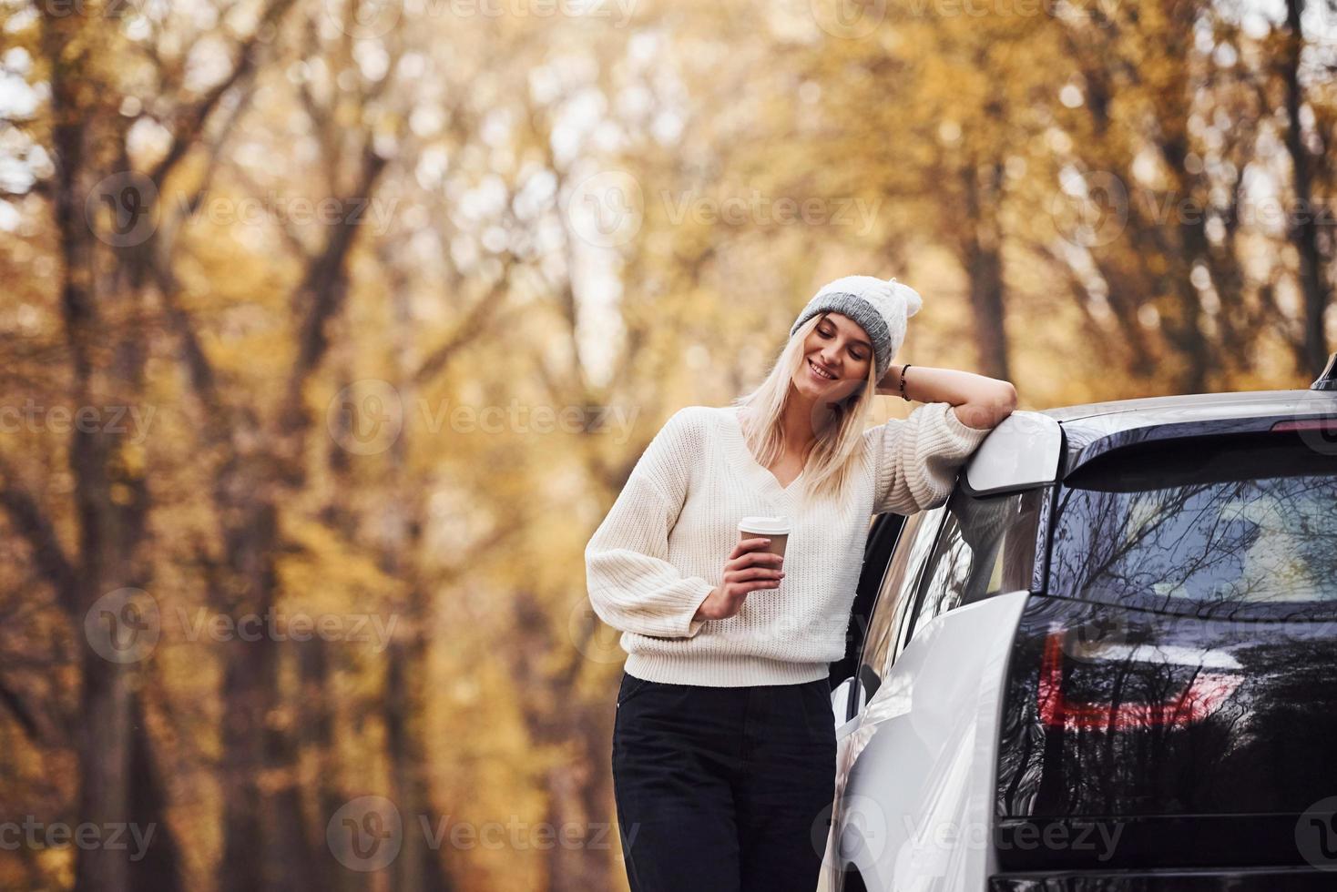 With cup of drink in hands. Girl have autumn trip by car. Modern brand new automobile in the forest photo