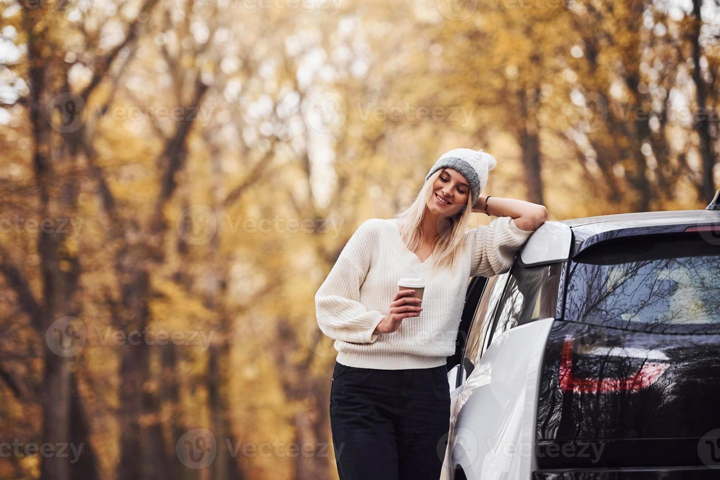 con taza de bebida en las manos. chica tiene viaje de otoño en coche. automóvil nuevo y moderno en el bosque foto