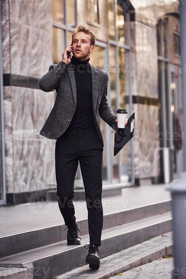 Man in elegant formal wear with cup of drink and notepad in hands is outside against modern building have a conversation by the phone photo