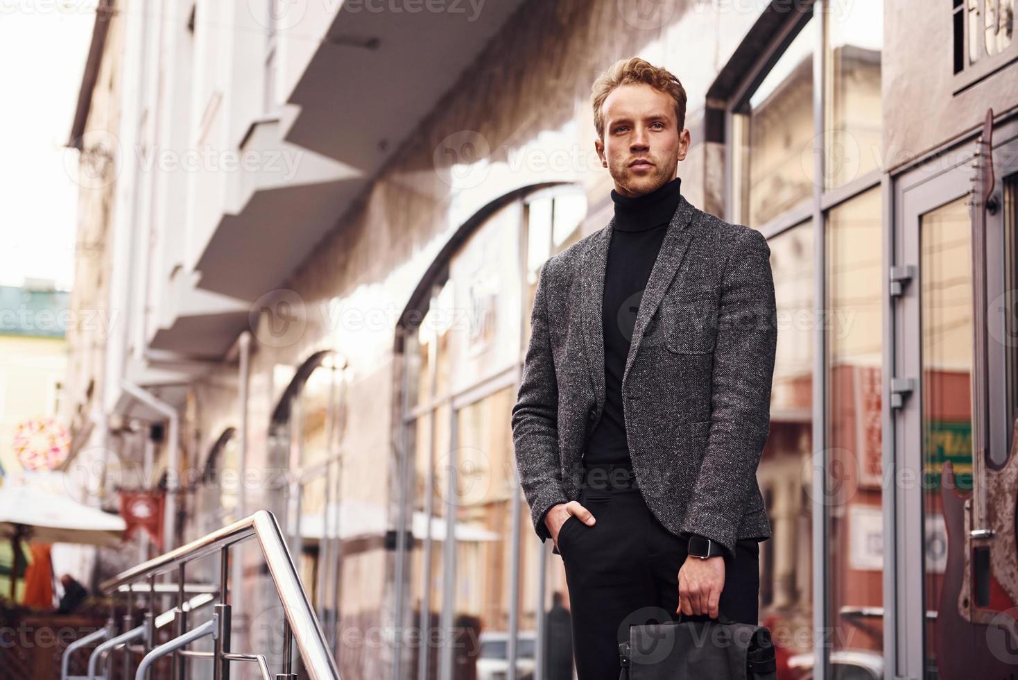 Man in elegant formal wear and with bag is outside against modern building photo