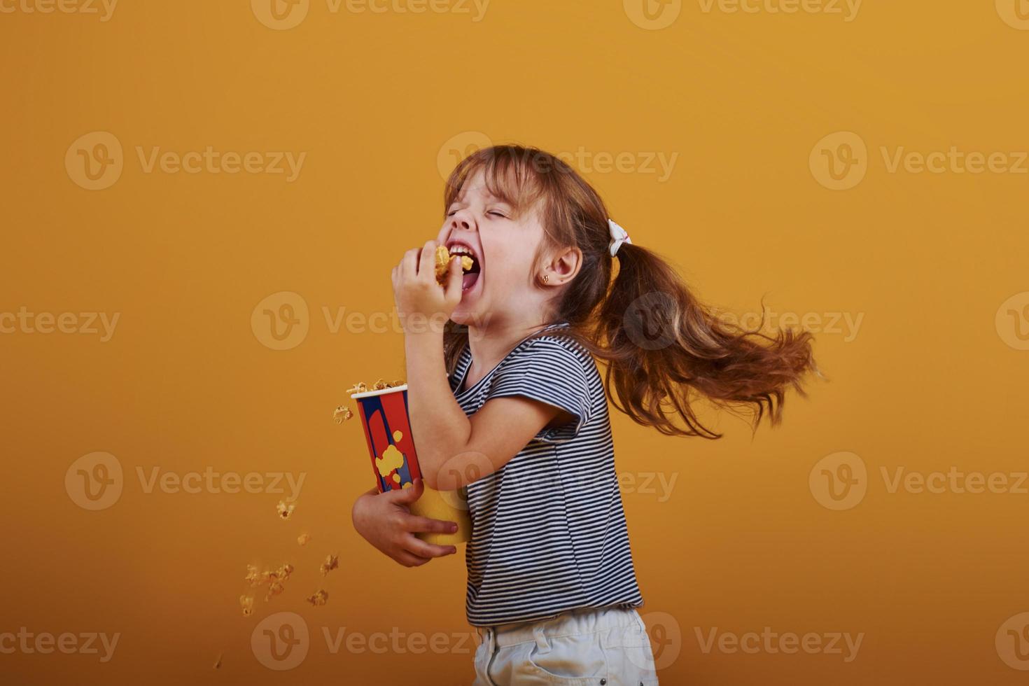 Cute little girl in in the studio eats popcorn and stands against yellow background photo