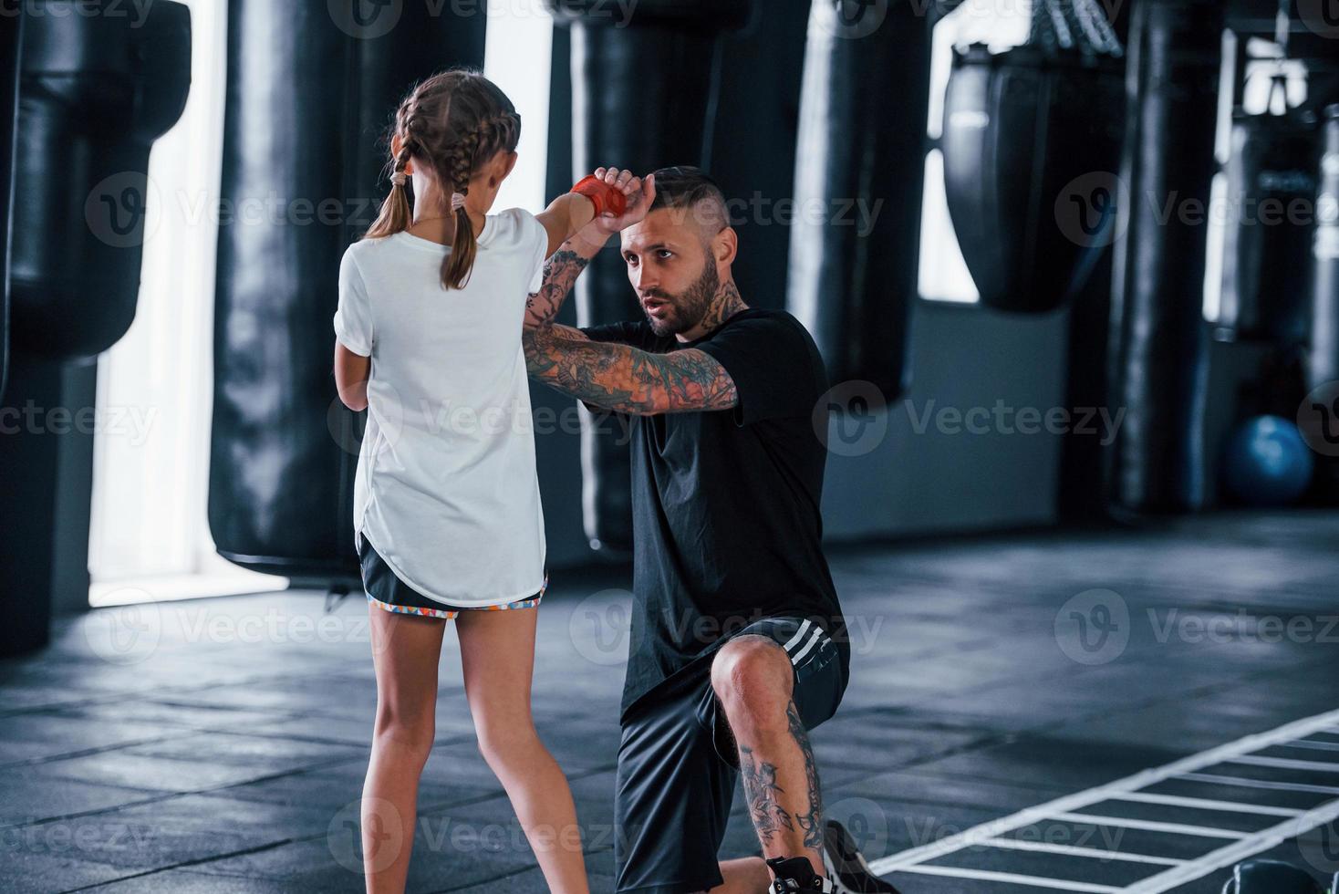 Basic punches. Young tattooed boxing coach teaches cute little girl in the gym photo