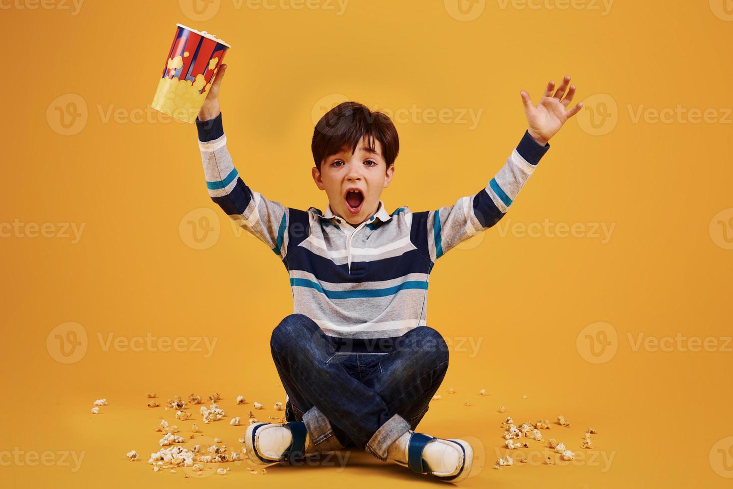 Cute little boy sitting and watching movie and eats popcorn against yellow wall photo