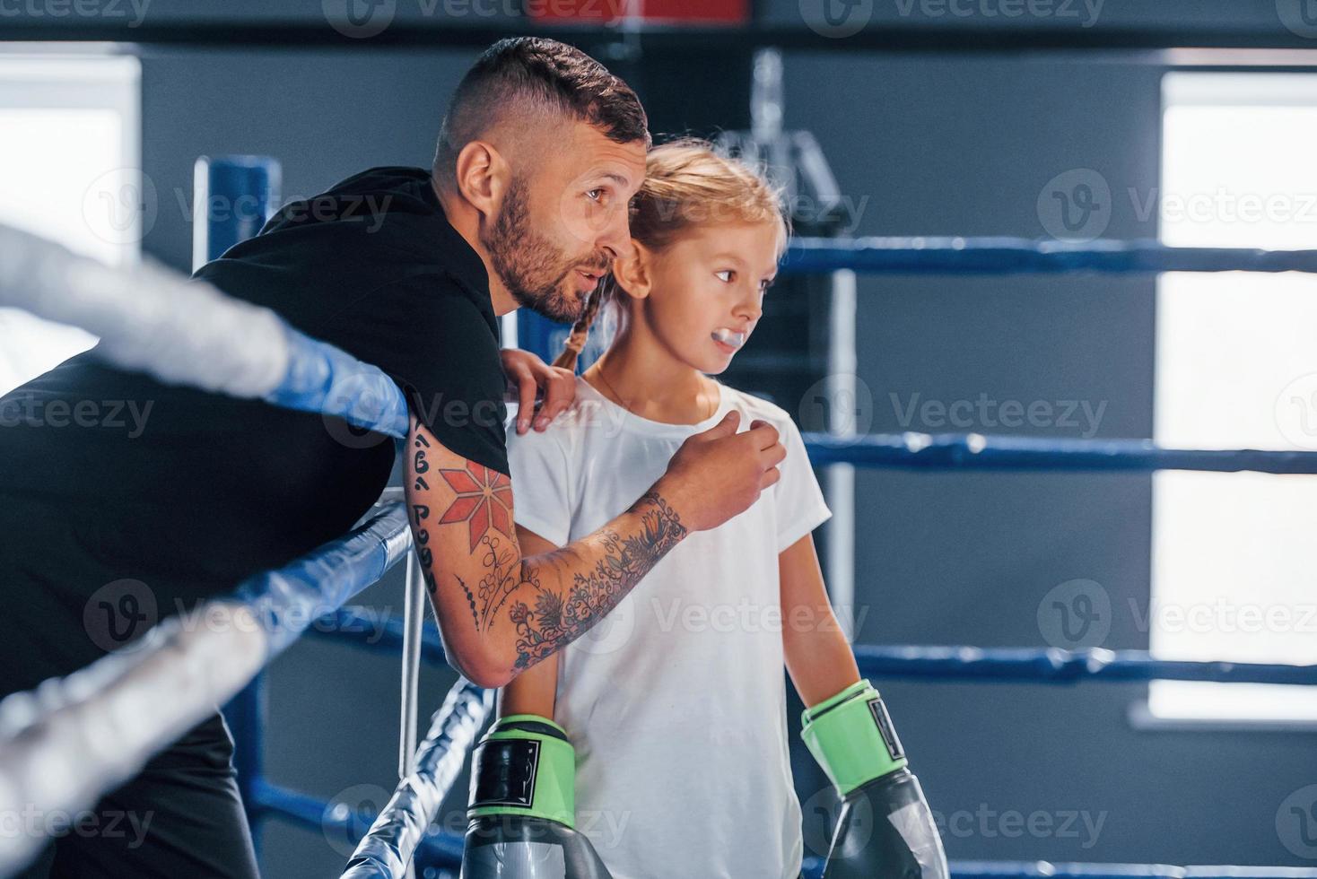 de pie en el ring de boxeo. joven entrenador de boxeo tatuado enseña a una linda niñita en el gimnasio foto