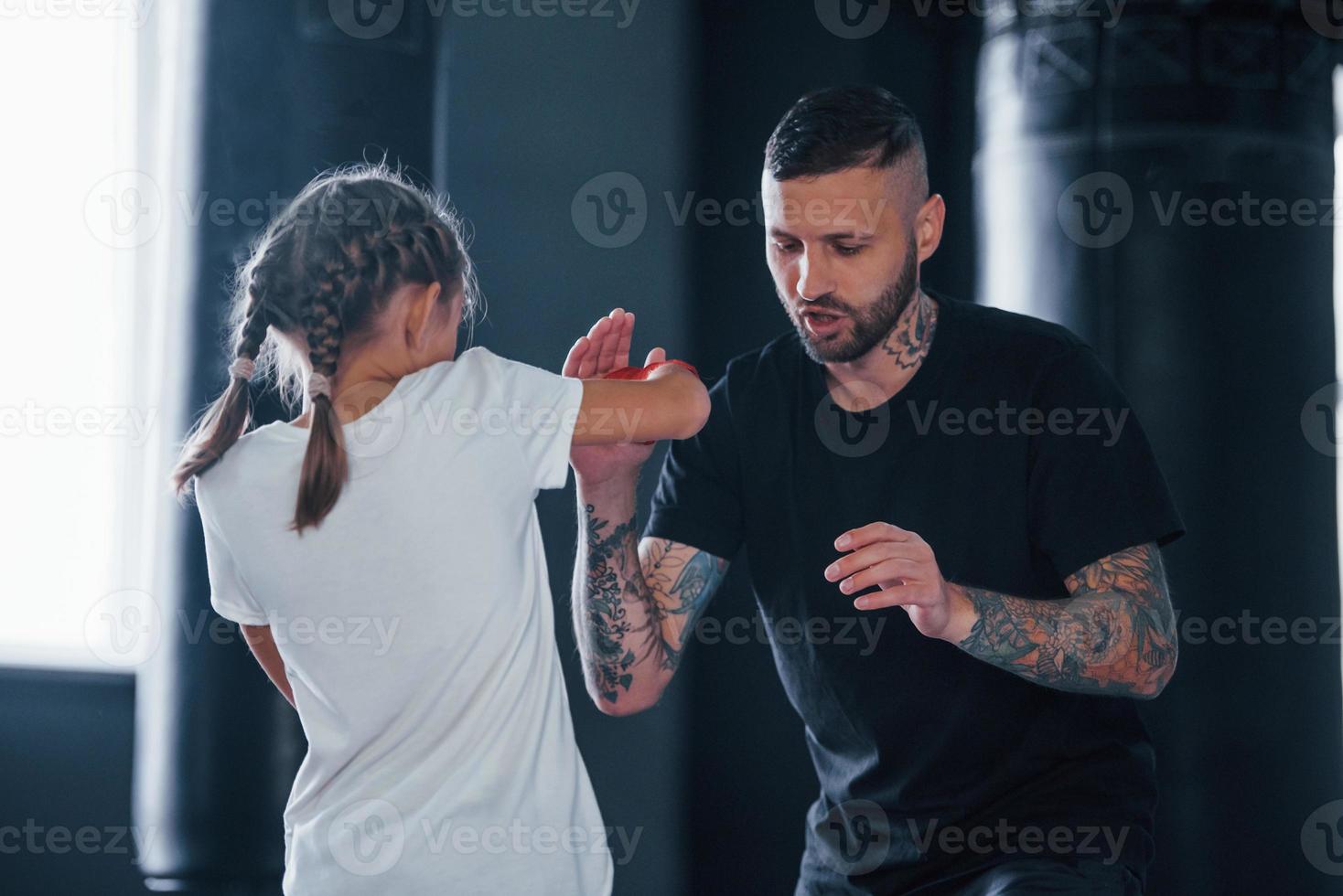 Basic punches. Young tattooed boxing coach teaches cute little girl in the gym photo