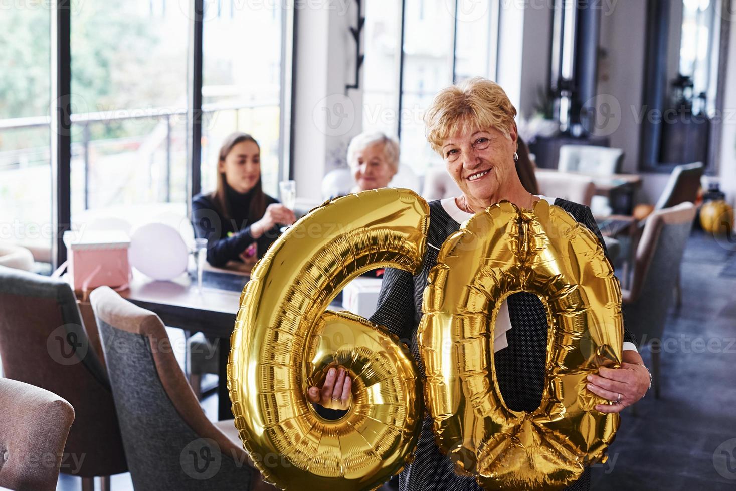 With balloons of number 60 in hands. Senior woman with family and friends celebrating a birthday indoors photo