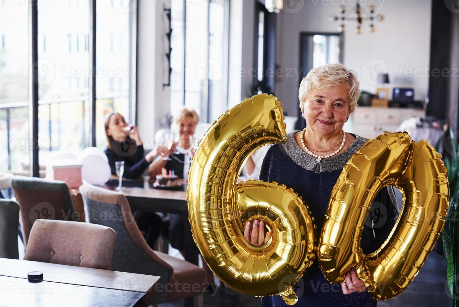 With balloons of number 60 in hands. Senior woman with family and friends celebrating a birthday indoors photo