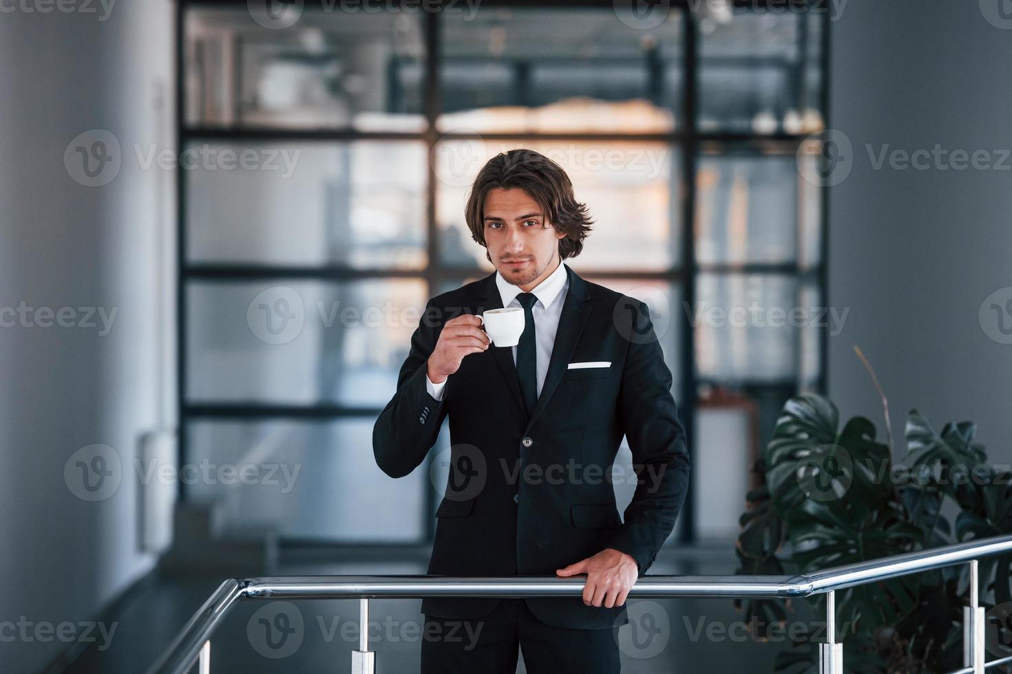Portrait of handsome young businessman in black suit and tie stands indoors with cup of drink photo