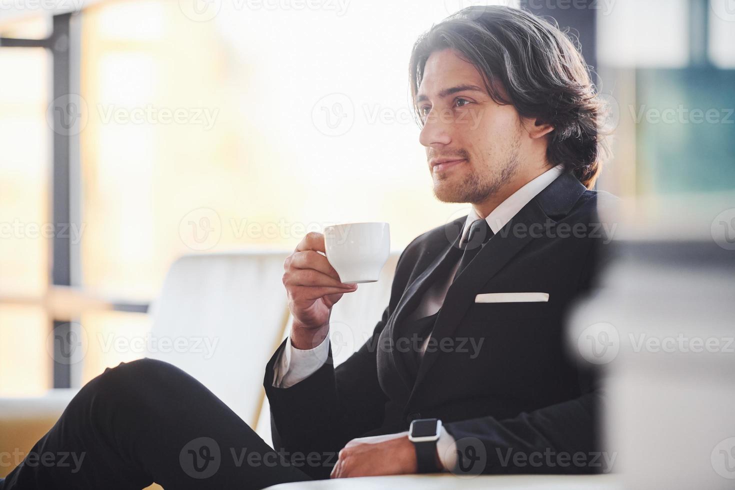 se sienta en el sofá con una taza de bebida. retrato de un apuesto joven hombre de negocios con traje negro y corbata foto