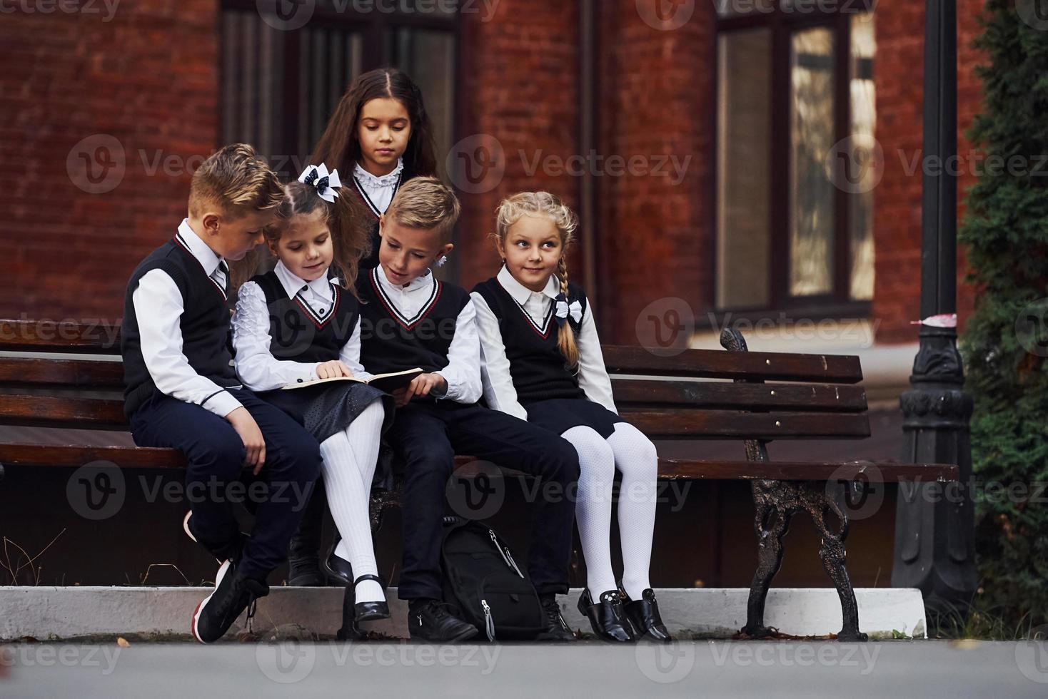 niños de la escuela en uniforme que se sienta al aire libre en el banco con bloc de notas foto