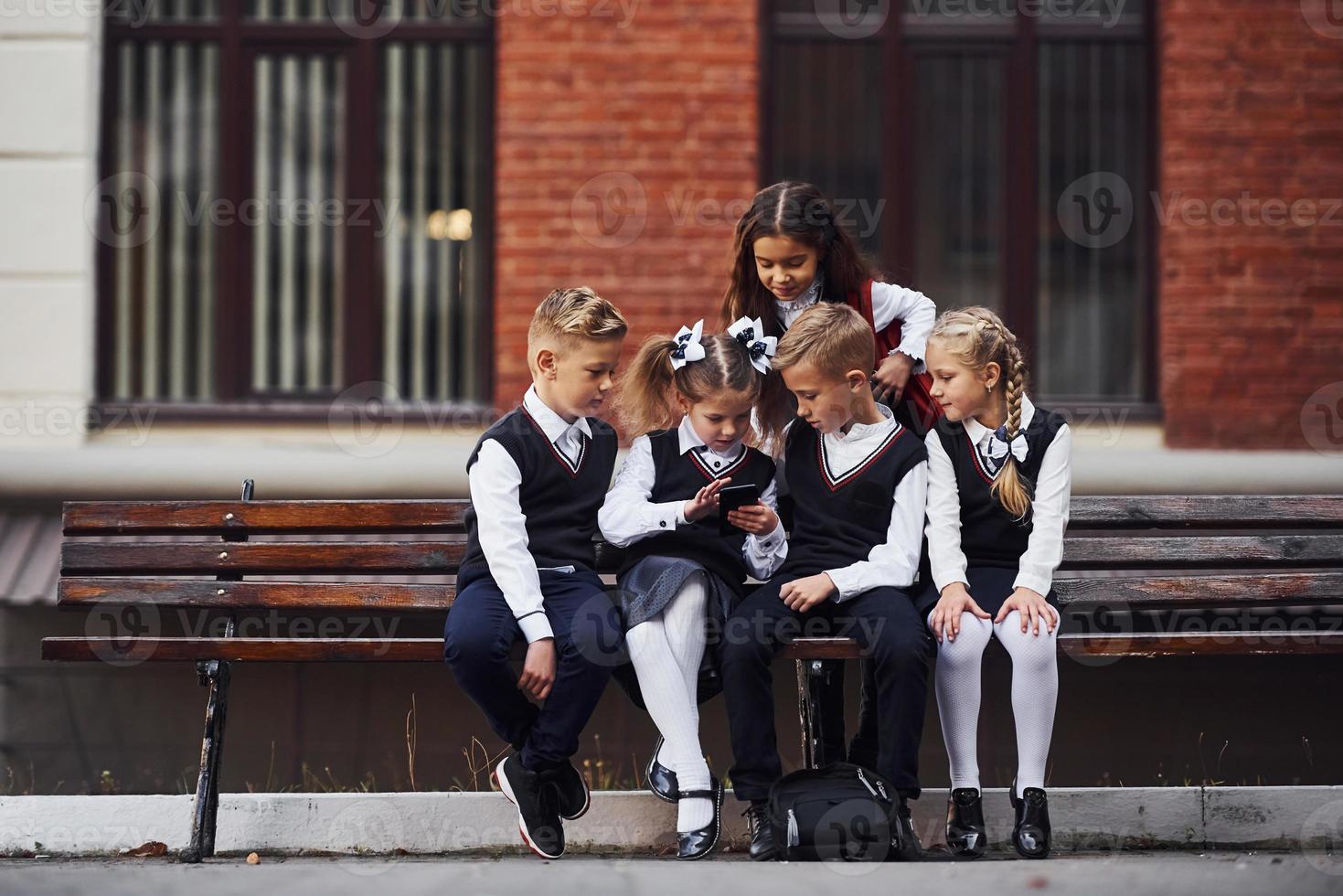 School kids in uniform that sits outdoors on the bench with smartphone photo