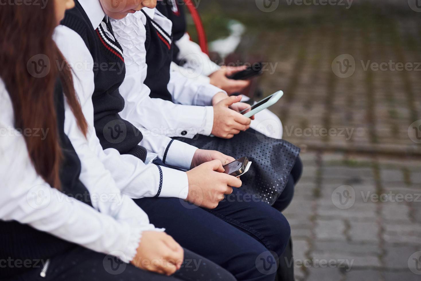 Close up view of school kids in uniform that sits on the bench with smartphones photo