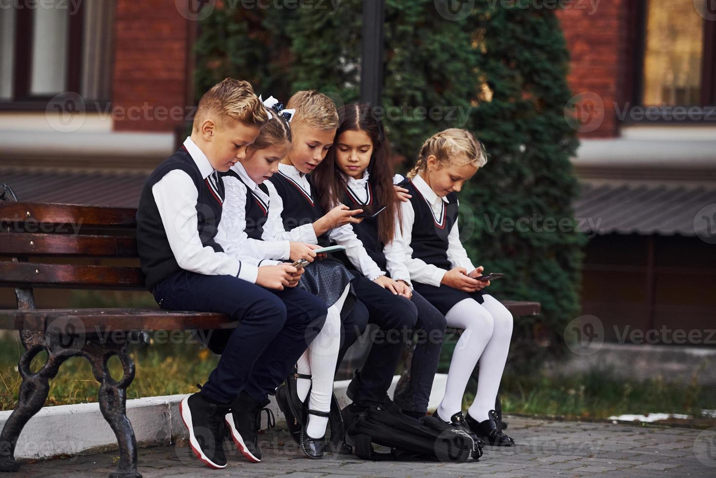 niños de la escuela en uniforme que se sienta al aire libre en el banco con teléfonos inteligentes foto