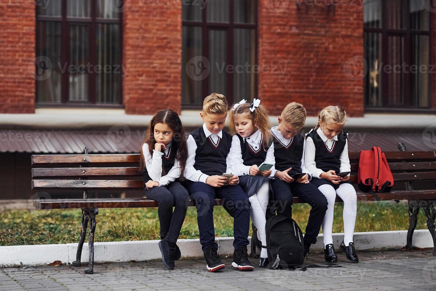 Group of kids in school uniform sits on the bench outdoors together near education building photo