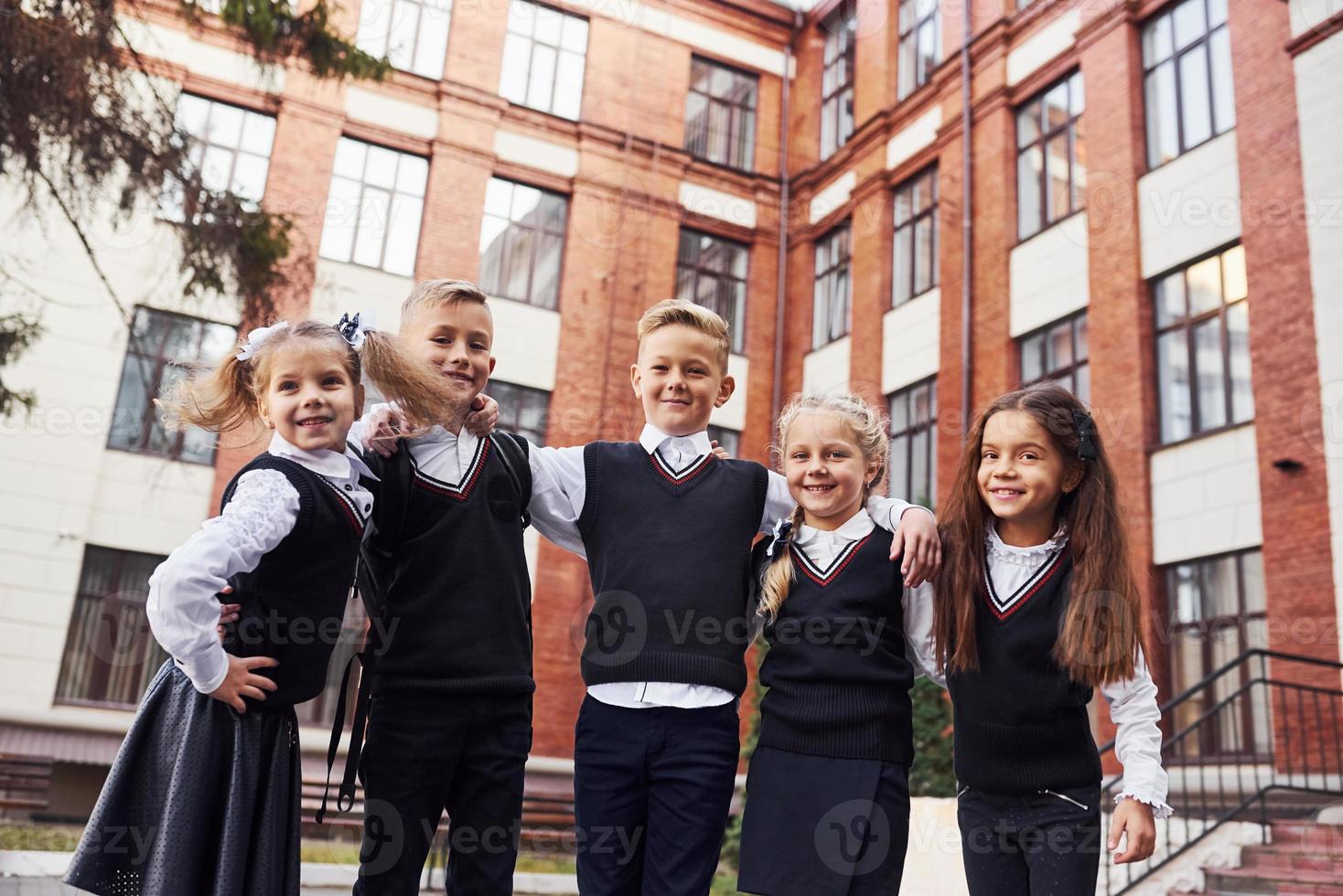 divirtiéndose y abrazándose unos a otros. grupo de niños con uniforme escolar que están juntos al aire libre cerca del edificio de educación foto