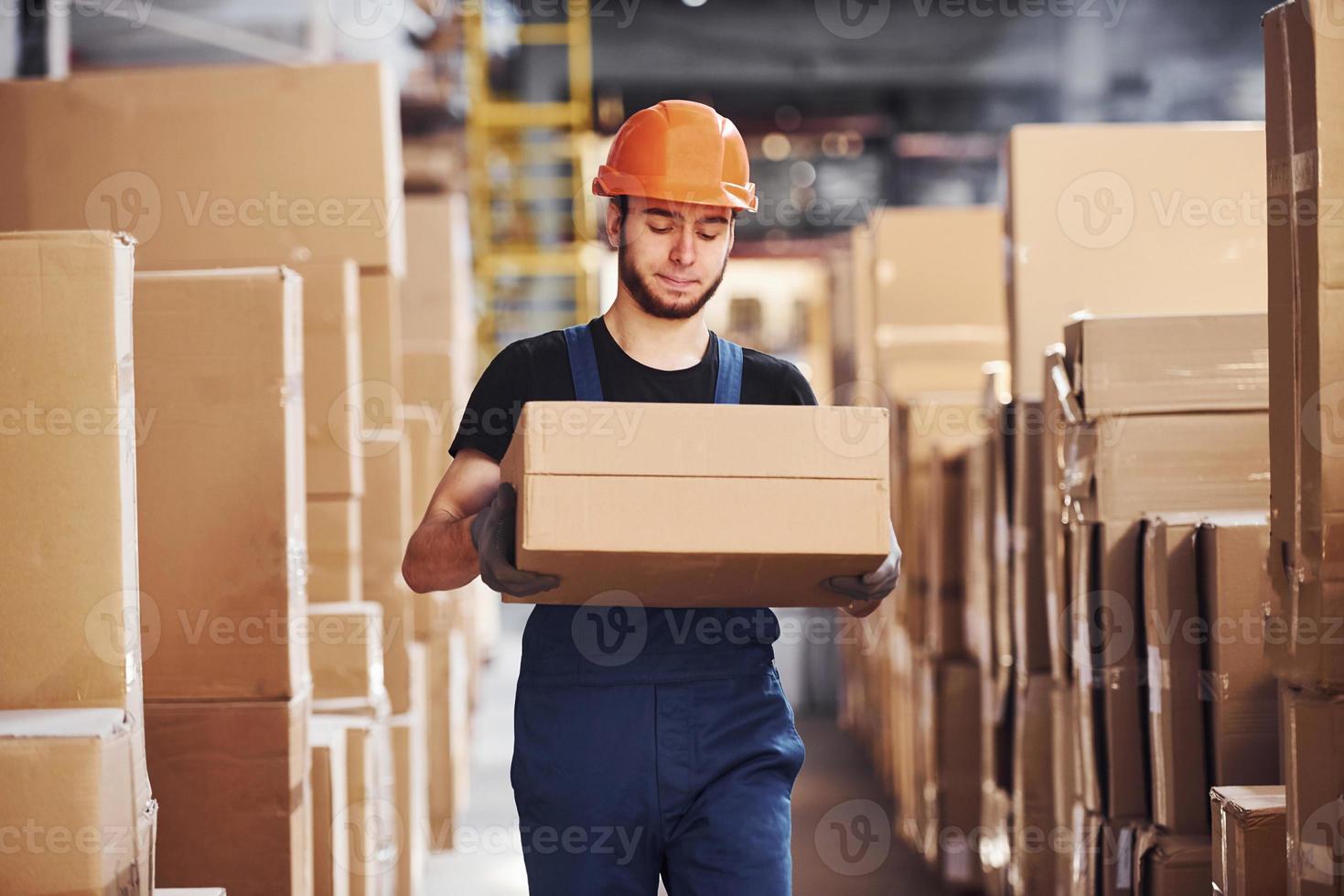 Young storage worker in uniform and hard hat carries box in hands photo