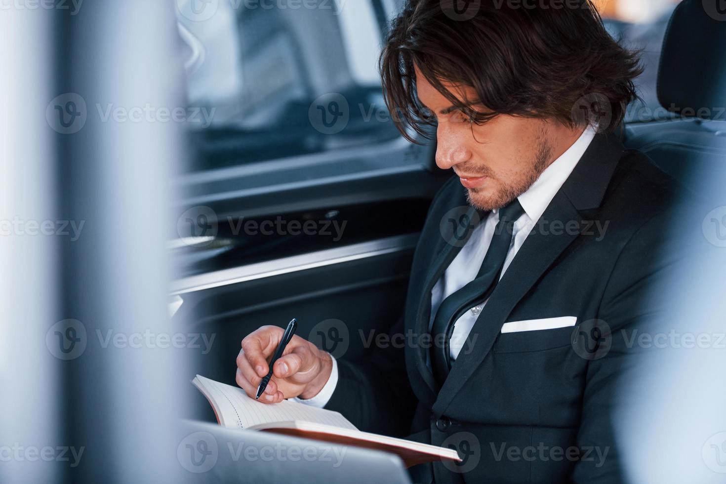 utilizando el bloc de notas. joven hombre de negocios con traje negro y corbata dentro de un automóvil moderno foto