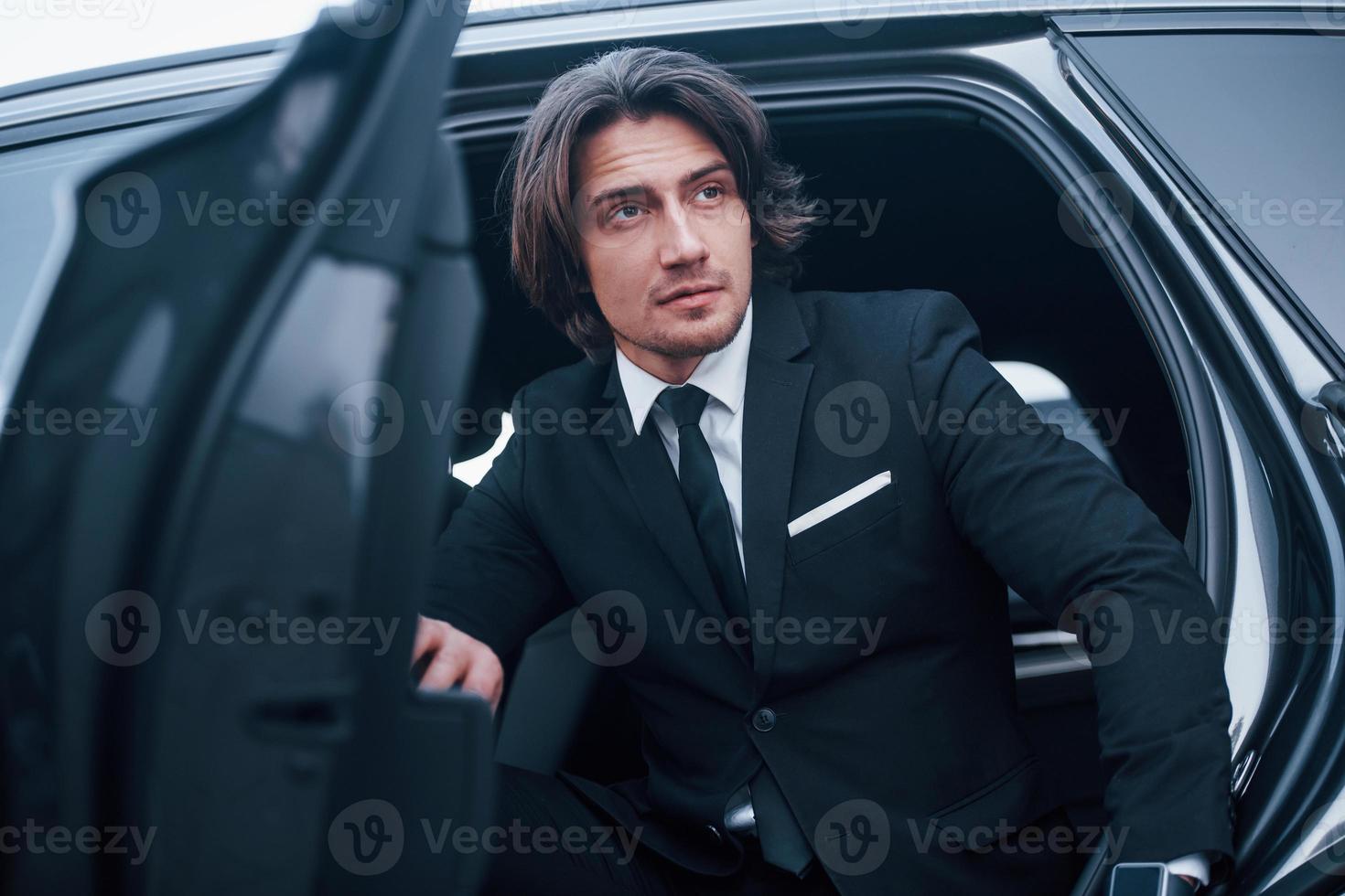 Portrait of handsome young businessman in black suit and tie walks out from modern car photo