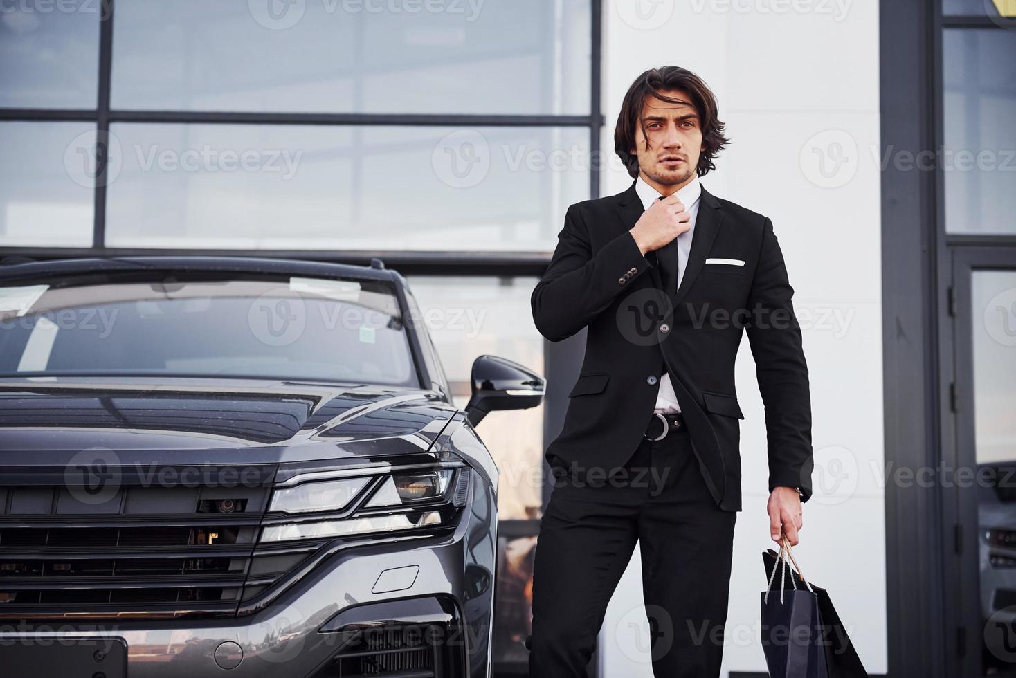 Portrait of handsome young businessman in black suit and tie outdoors near modern car and with shopping bags photo