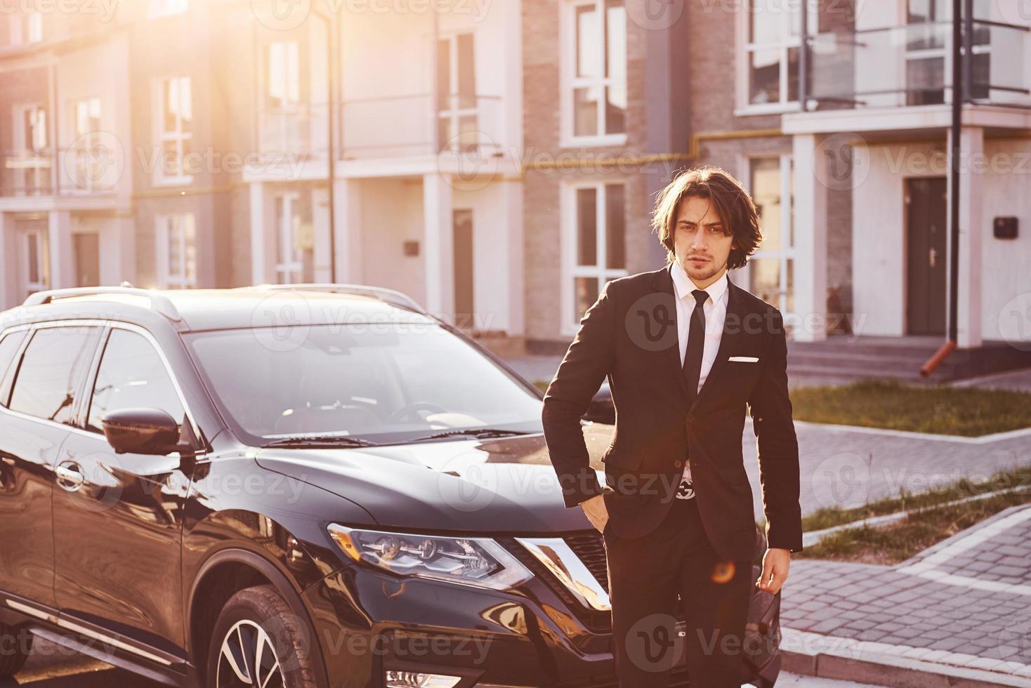 Portrait of handsome young businessman in black suit and tie outdoors near modern car in the city photo