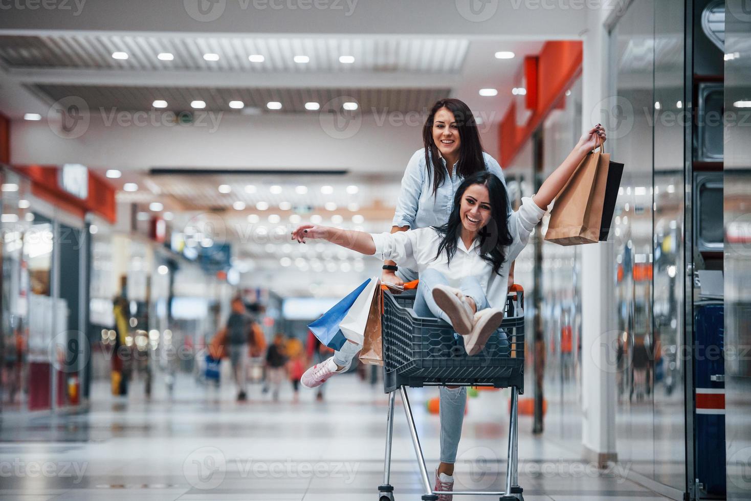 dos mujeres jóvenes se divierten corriendo y montando en la cesta de la compra en el supermercado foto