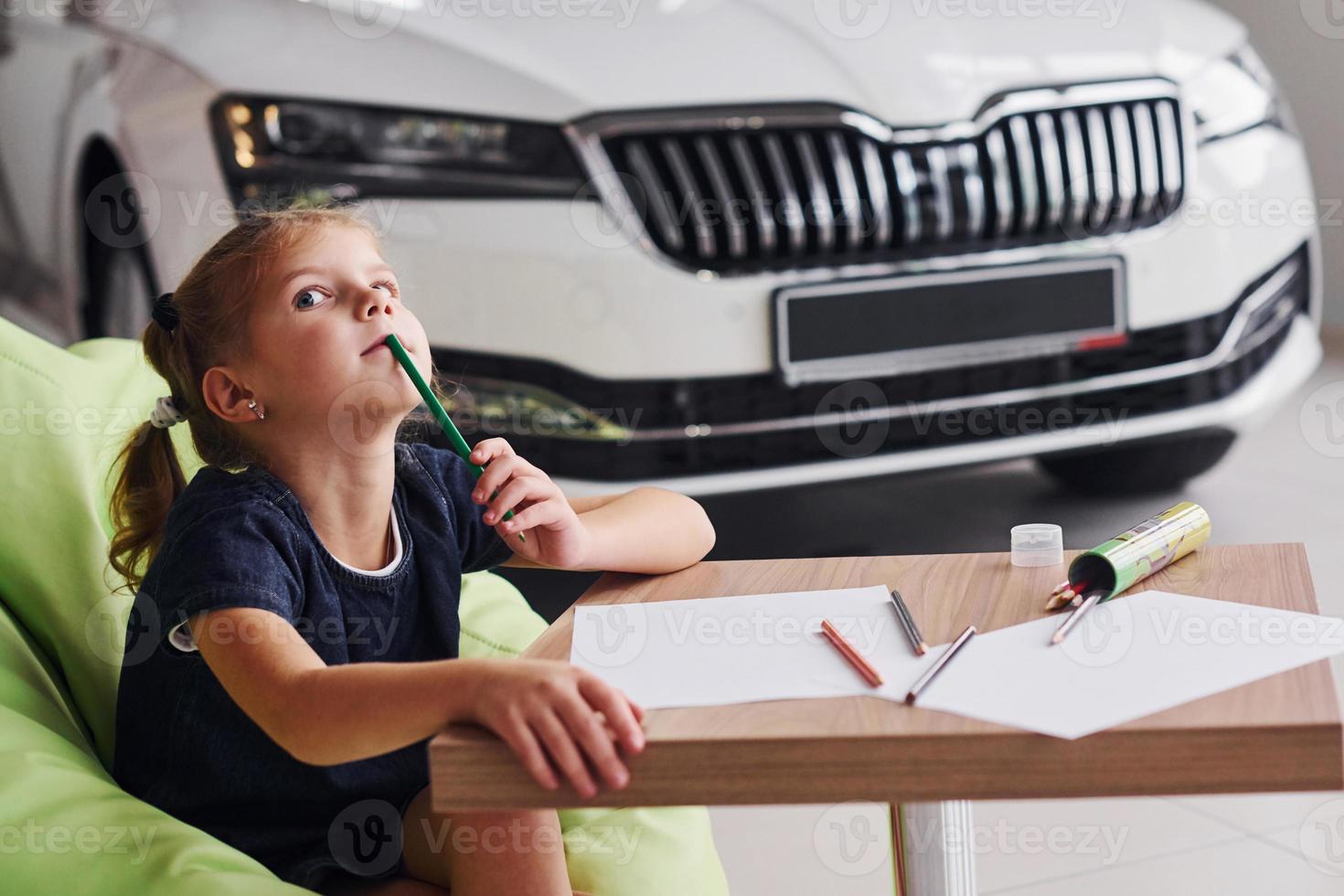 Cute little girl sits on the soft green chair by the table with pencil and paper sheets. Near modern automobile photo