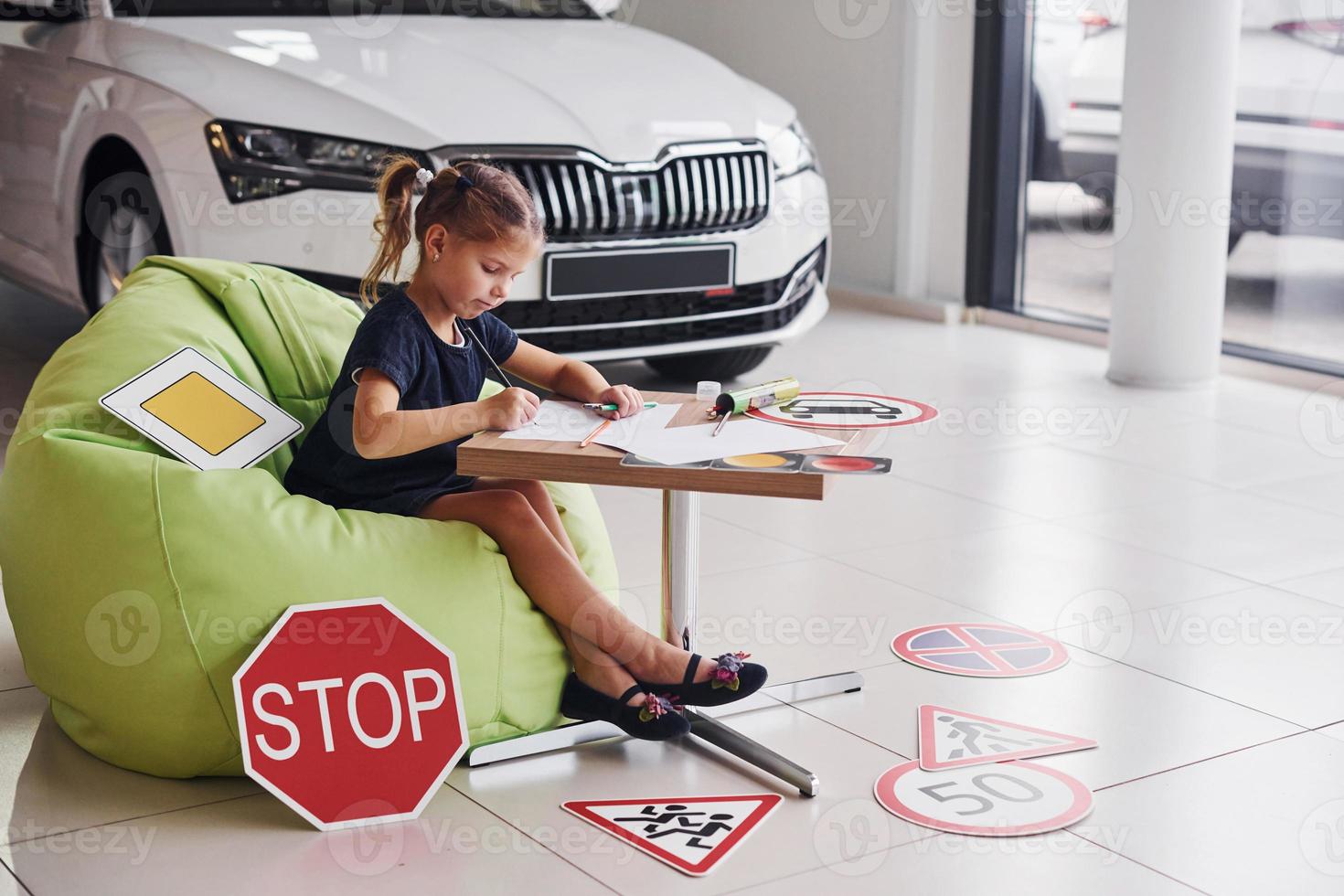 Cute little girl sits on the soft green chair by the table with pencil and paper sheets. Near modern automobile and road signs on the floor photo