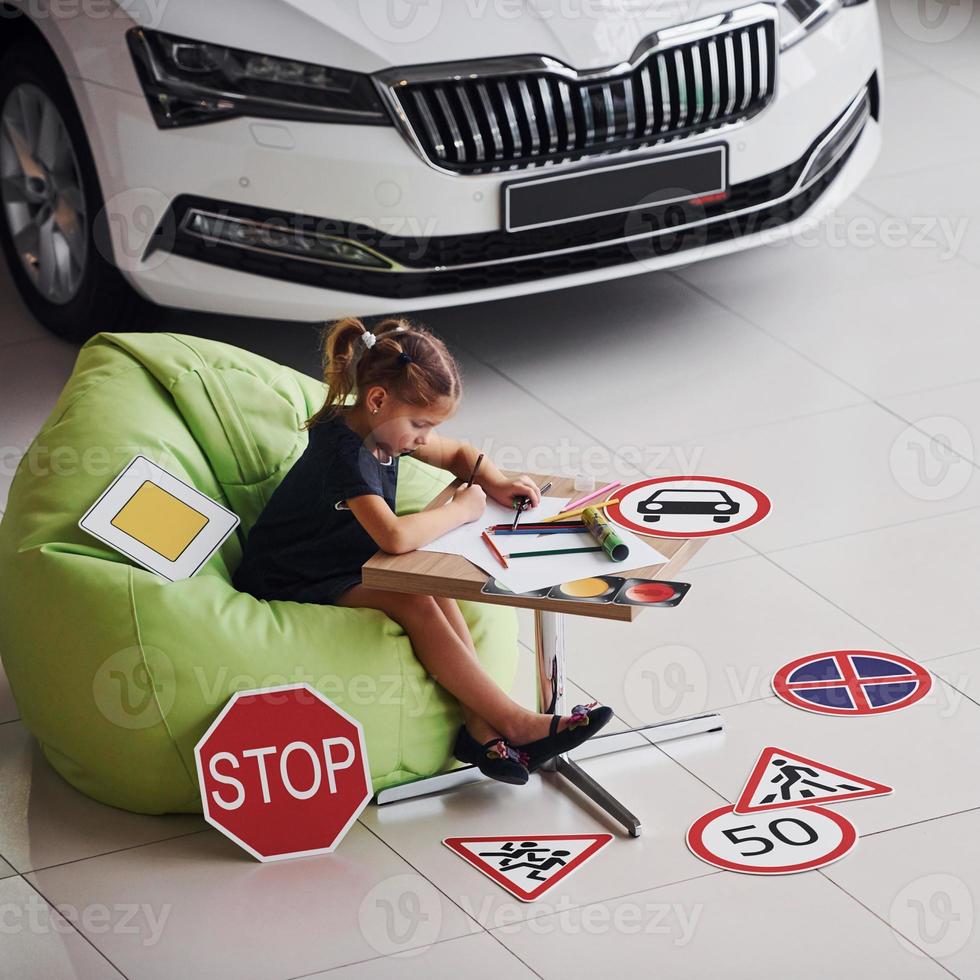 Cute little girl sits on the soft green chair by the table with pencil and paper sheets. Near modern automobile and road signs on the floor photo