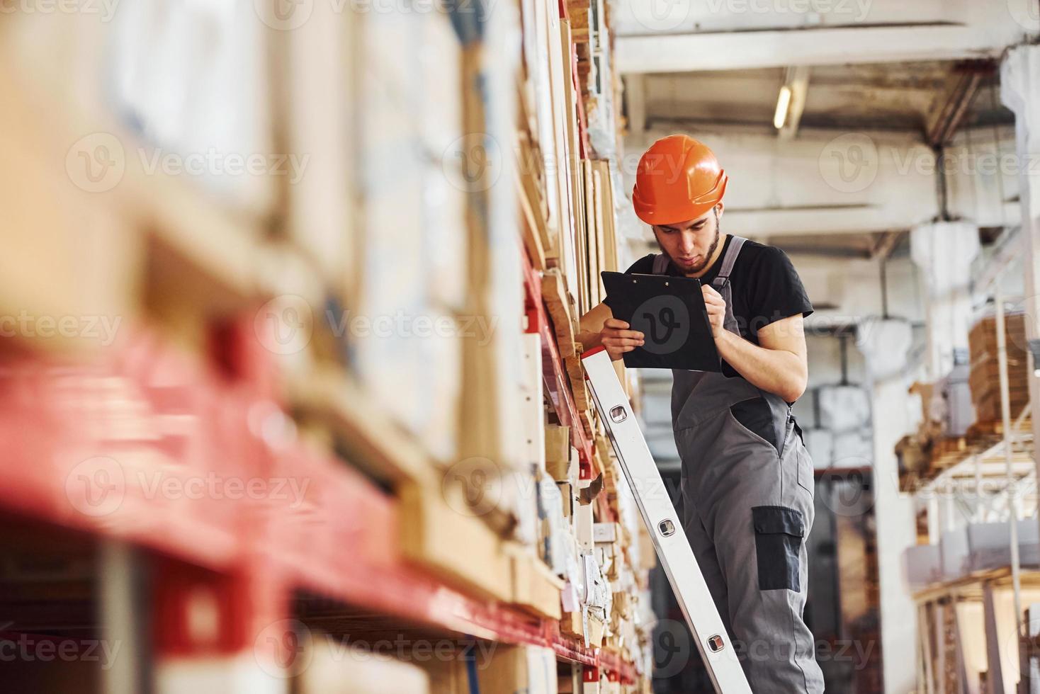 Storage worker stands on the ladder in uniform and notepad in hands and checks production photo