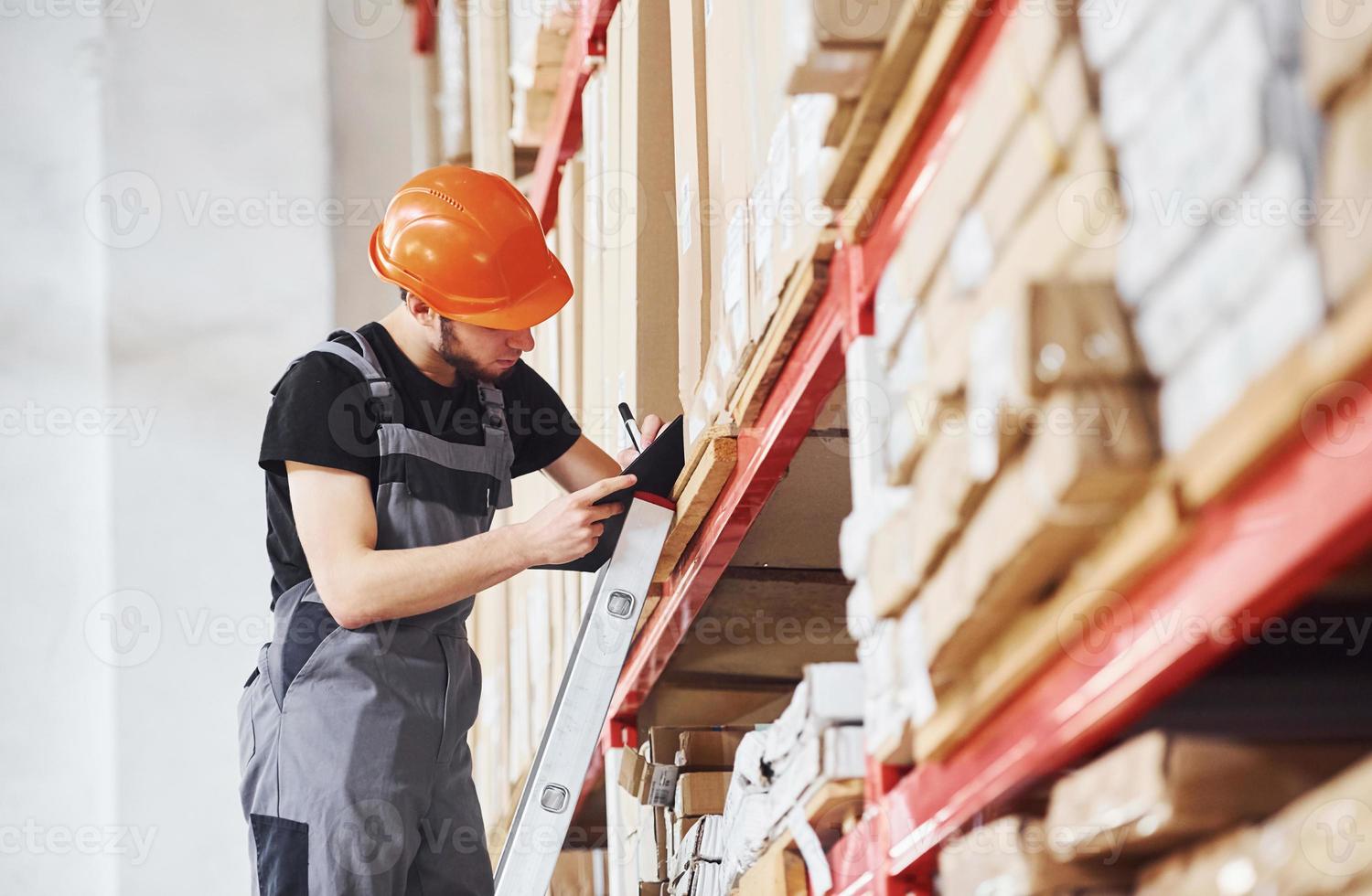 Storage worker stands on the ladder in uniform and notepad in hands and checks production photo