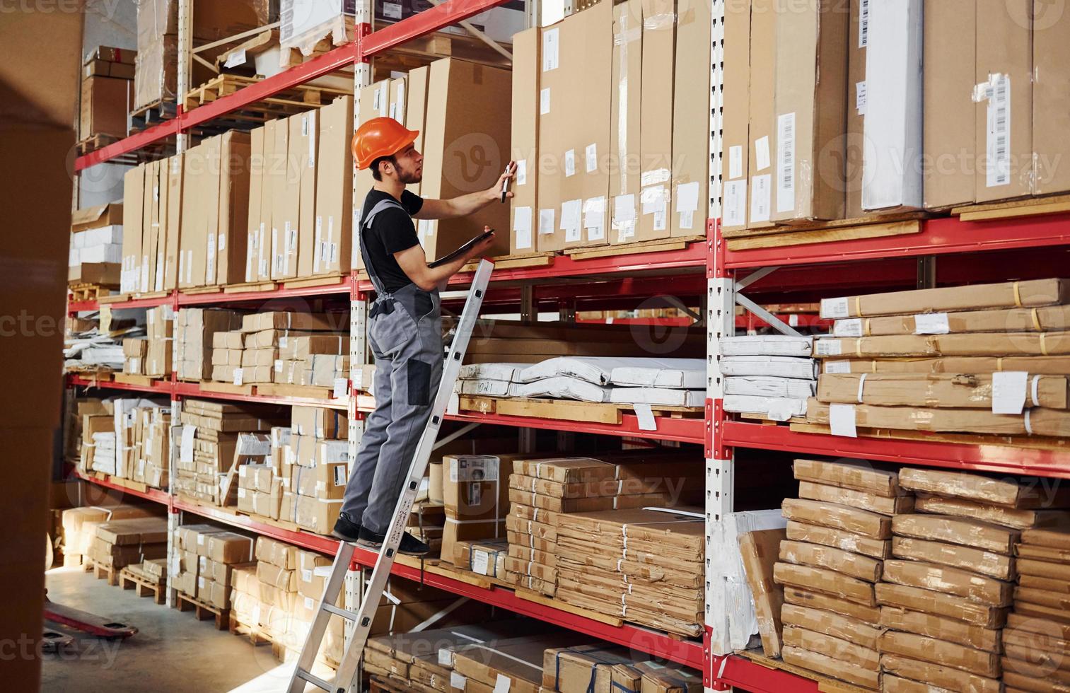 Storage worker stands on the ladder in uniform and notepad in hands and checks production photo