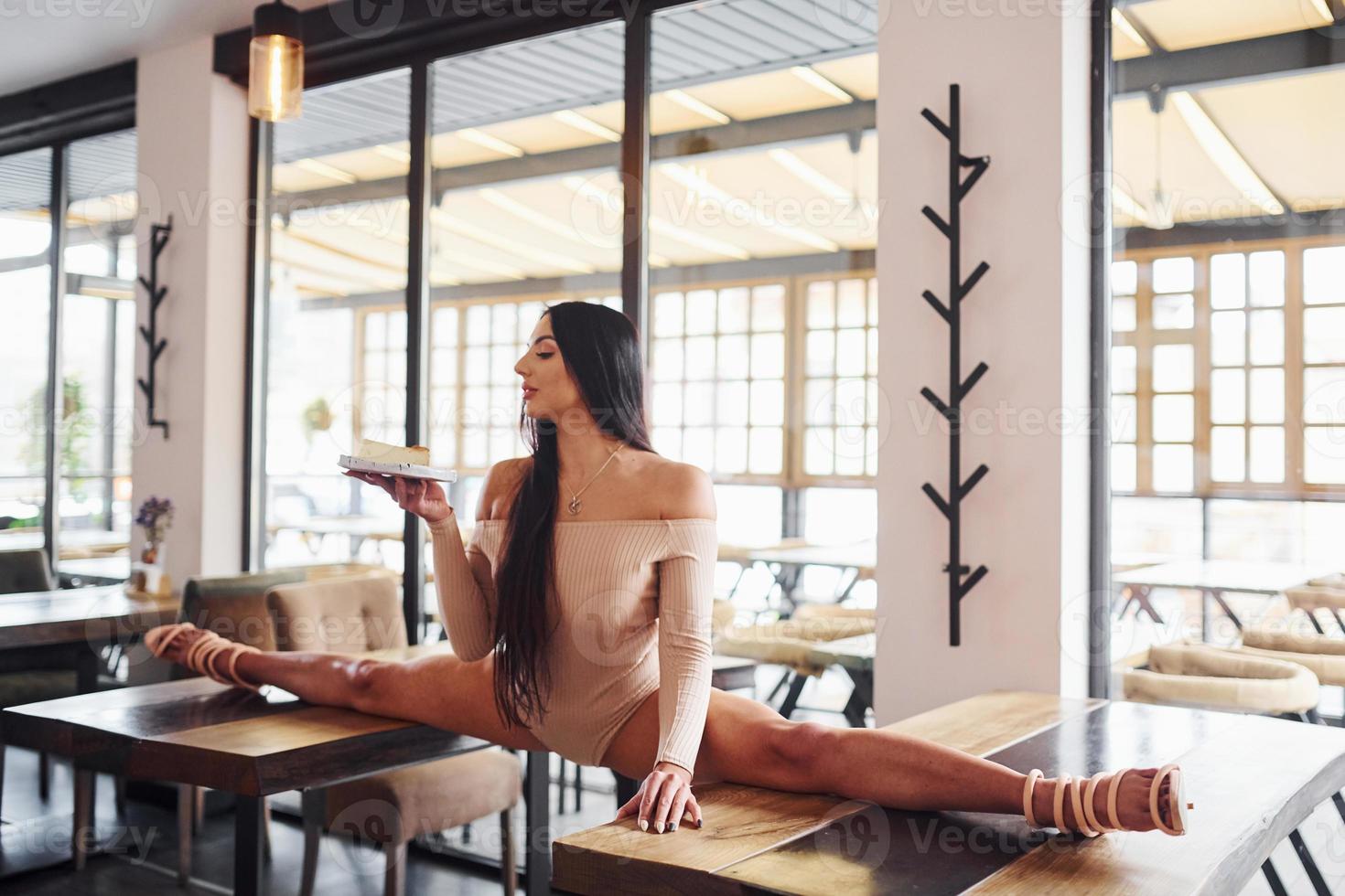 Young brunette in sexy wear soing stretches by sitting on the table and holds plate with food in restaurant photo