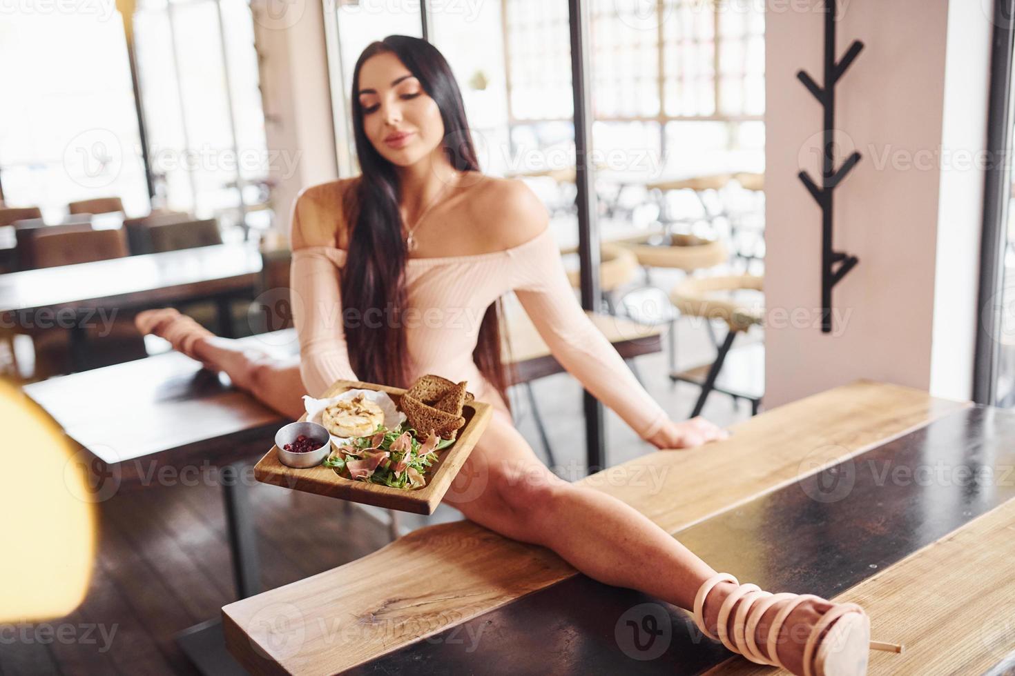 Young brunette in sexy wear soing stretches by sitting on the table and holds plate with food in restaurant photo