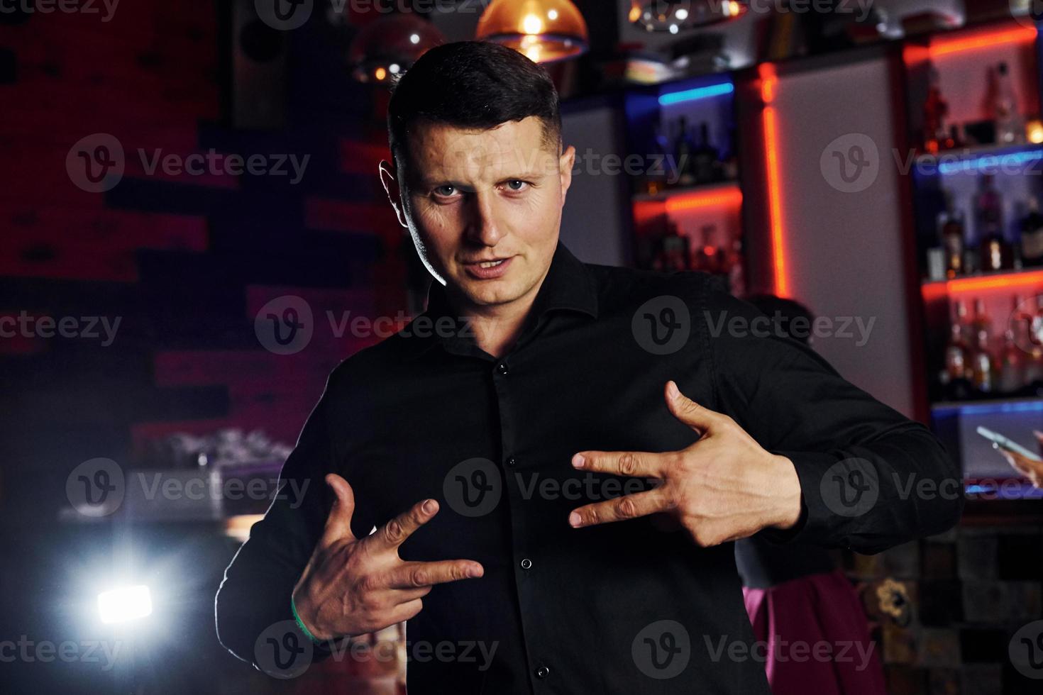 Man in black shirt stands inside of night club and posing for the camera photo